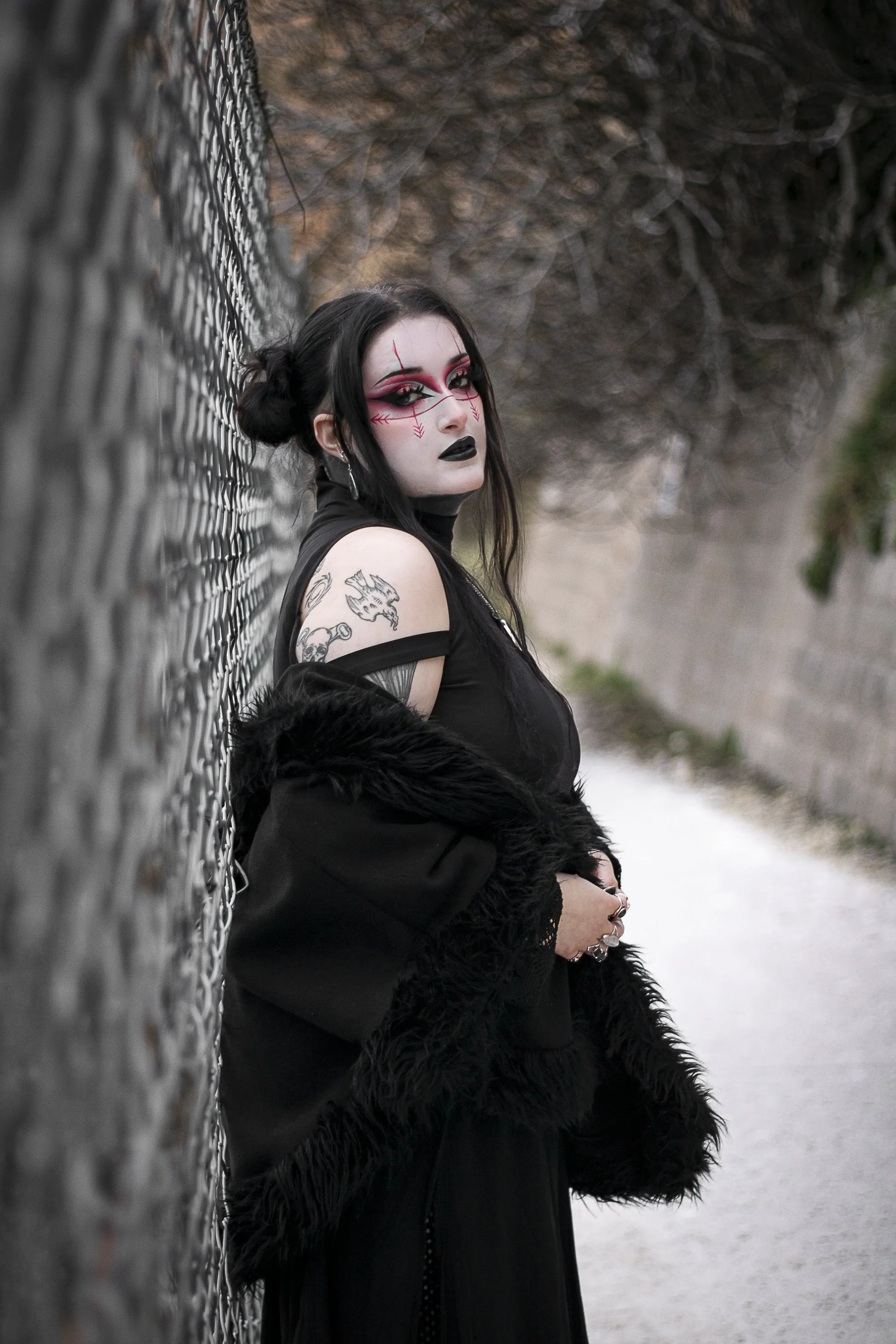 A woman with gothic makeup and tattoos leaning against a chain-link fence in an outdoor setting.