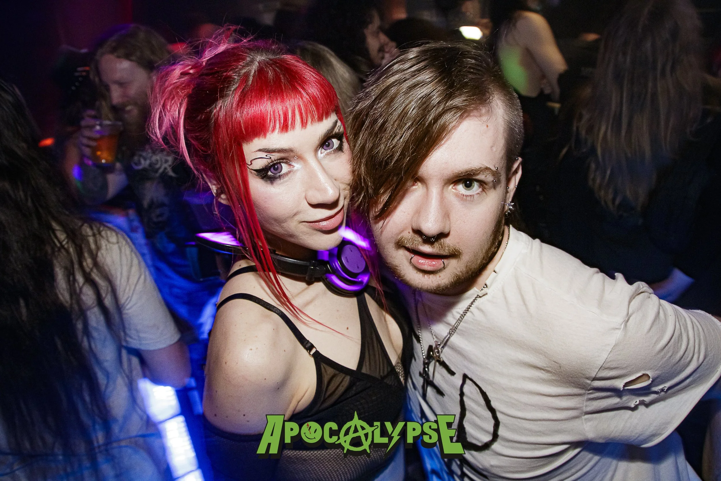 A young man and woman at a party or nightclub, crowd in the background, woman with bright red hair and gothic style makeup, wearing a black top with straps and purple headphones, man with brown hair, piercings, and a white torn shirt, both looking at
