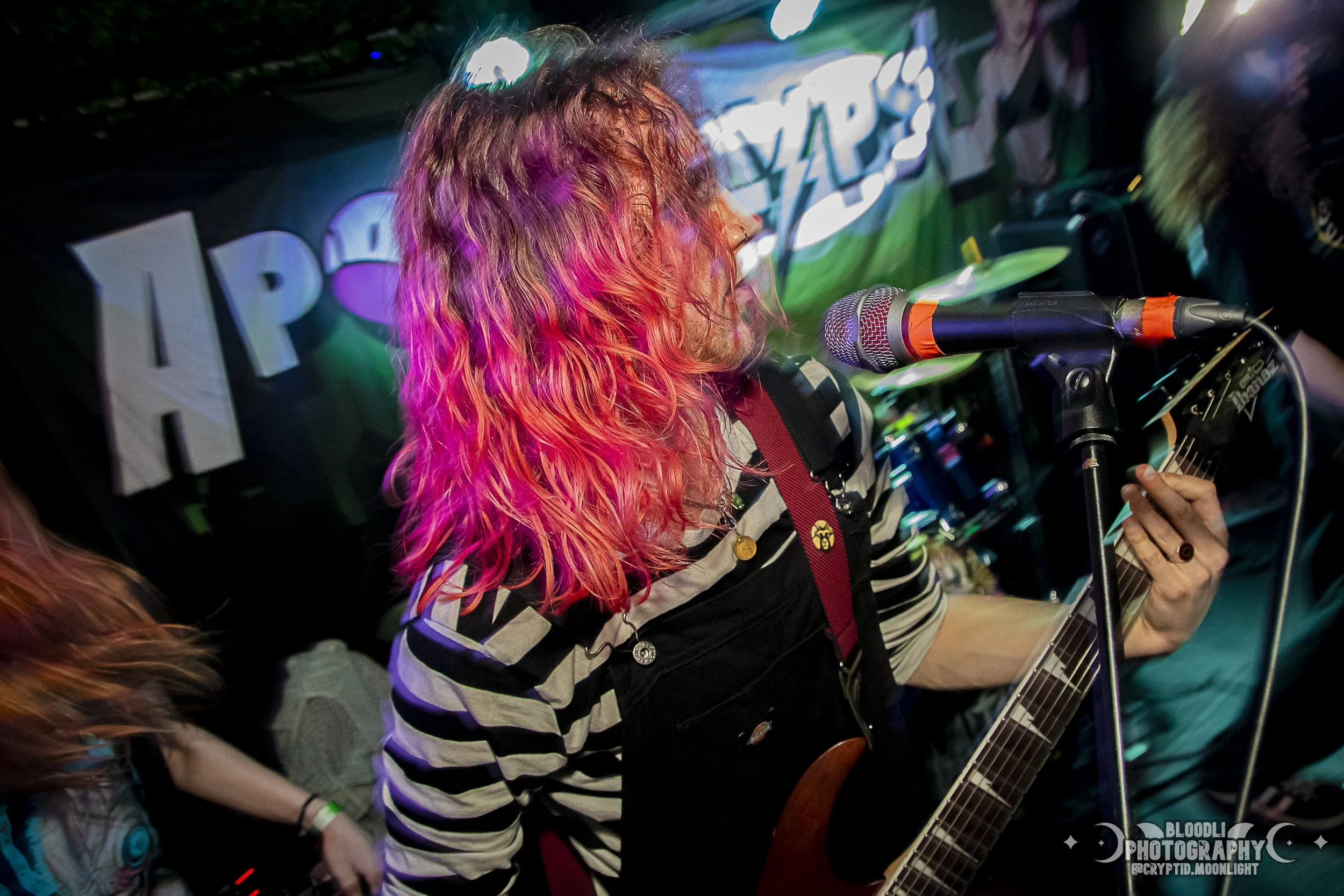 A woman with pink and orange hair singing and playing guitar at a microphone during a performance at a live music venue.
