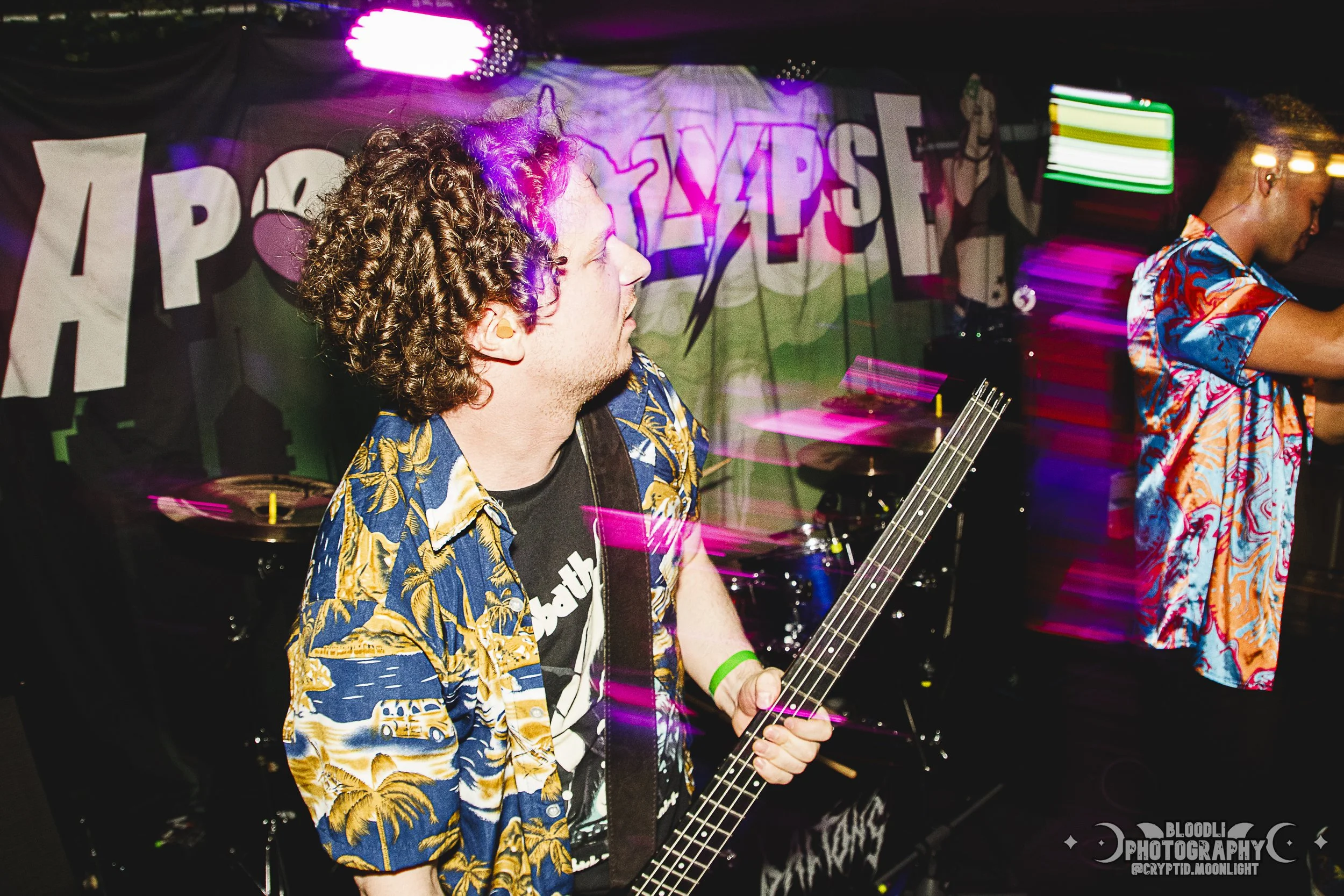 A musician with curly hair wearing a tropical shirt playing a bass guitar during a live performance at a club or concert venue with colorful lights and a signed backdrop in the background.