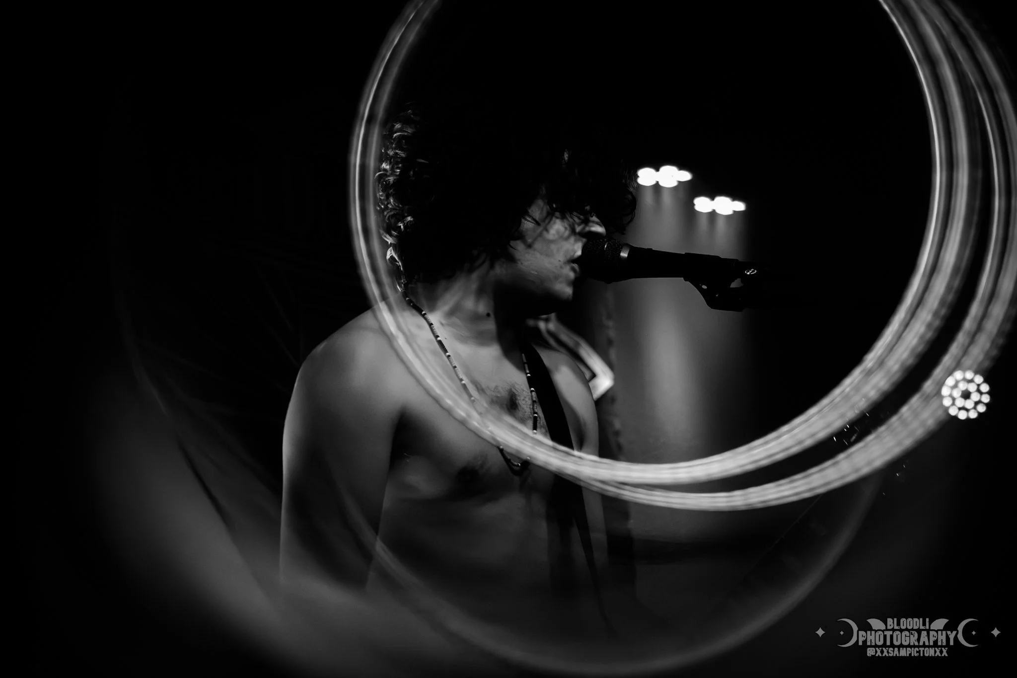 Black and white photo of a male musician with curly hair singing into a microphone on stage, viewed through a circular lighted mirror.