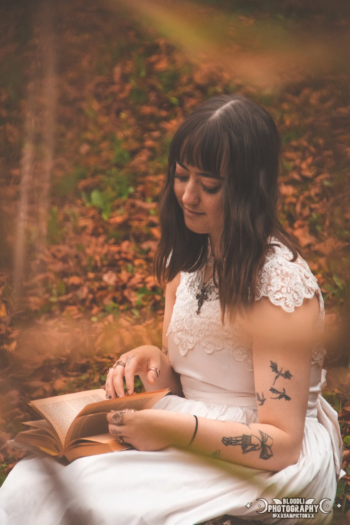 A young woman with dark hair and tattoos, wearing a white dress, sitting outdoors on a bed of fallen leaves, reading a book.