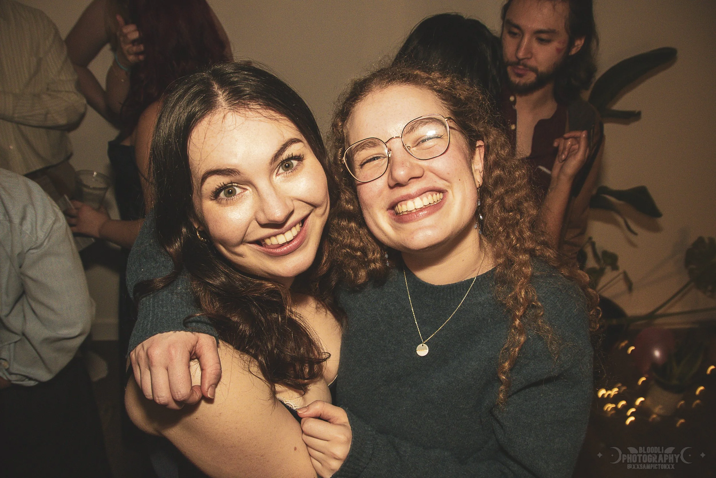 Two women smiling and hugging each other at a party, with other people in the background.