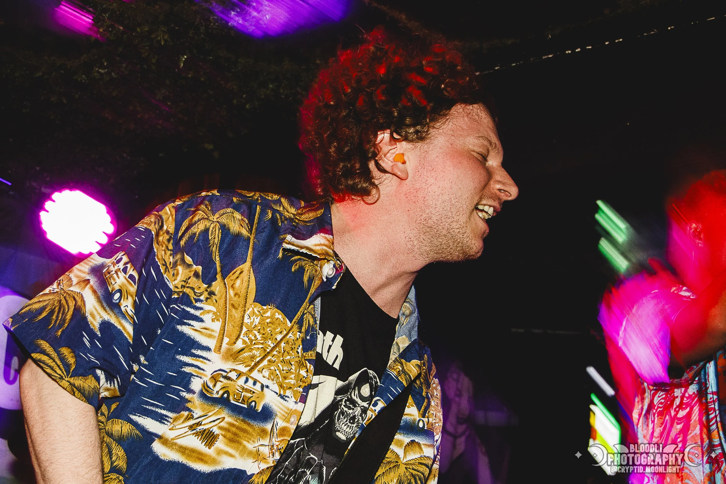 A man with curly red hair and light skin is smiling and dancing at a party, wearing a colorful Hawaiian shirt with palm trees and cars, under purple and green party lights.