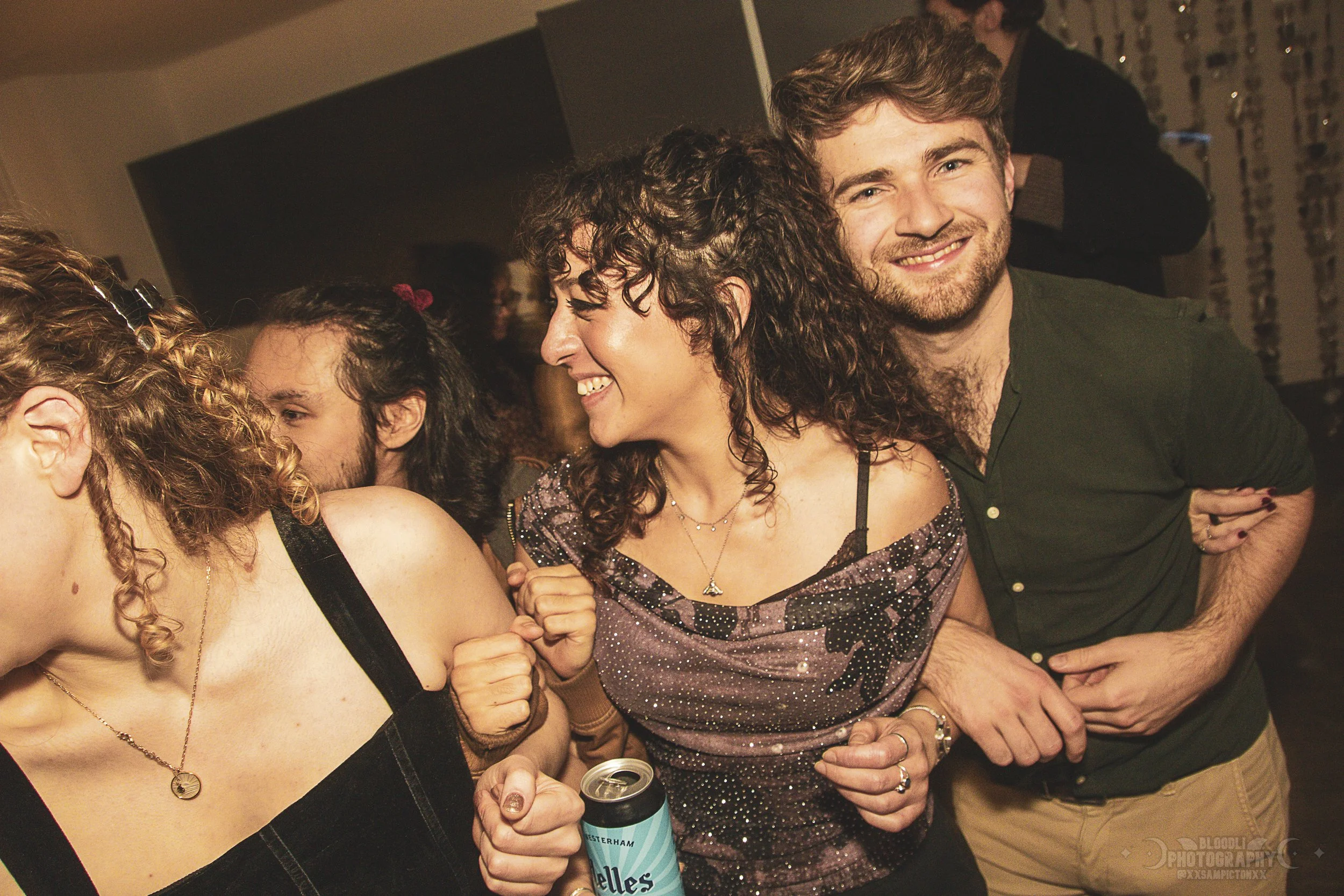 Group of people dancing and smiling at a party, with a woman holding a can of Yankee Elle in the foreground.