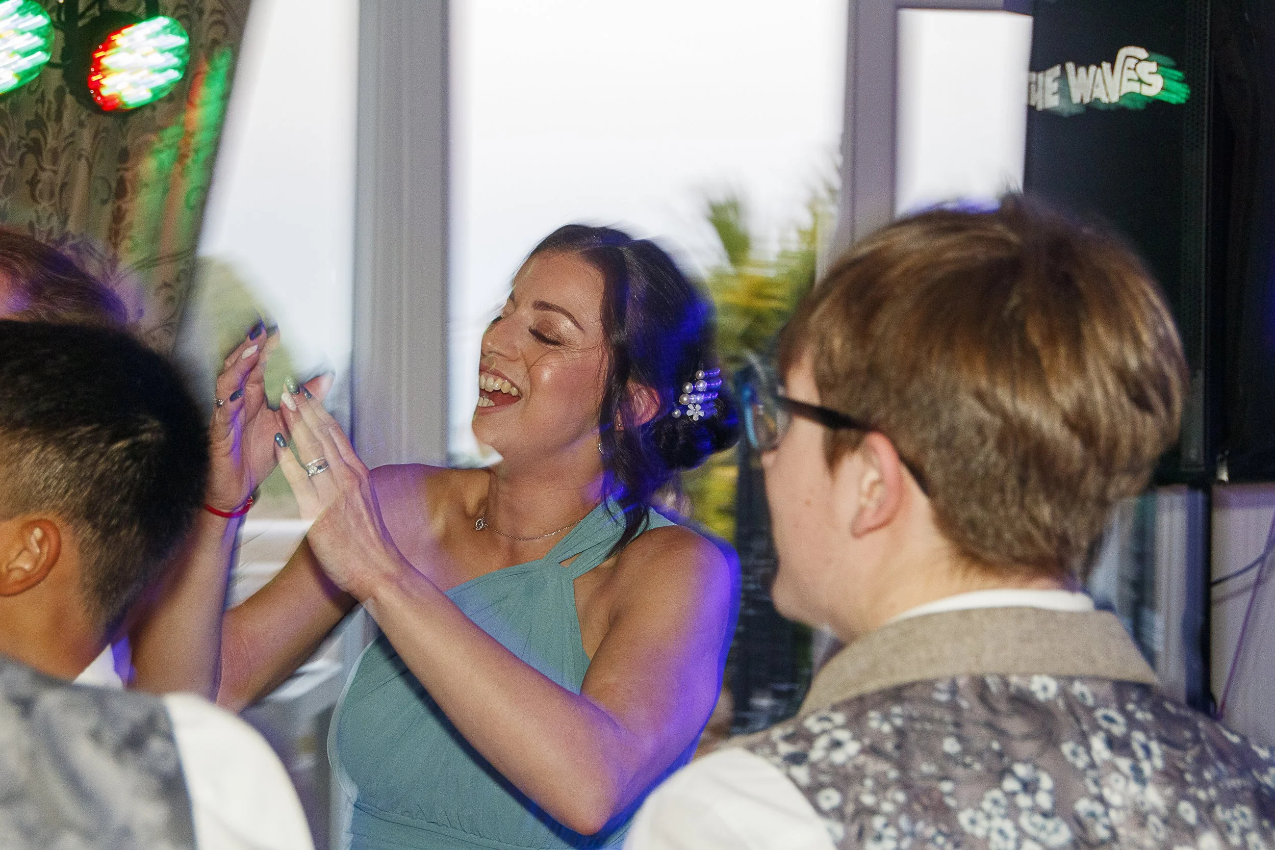 Group of people celebrating at a party, woman smiling and clapping, wearing a green dress, others in casual attire, decorations with balloons in the background.