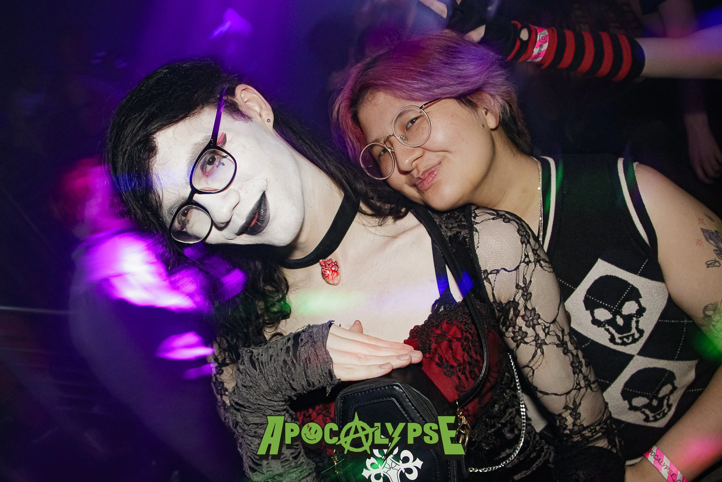 Two women at a party, one with dark hair, glasses, and goth makeup, the other with purple hair and glasses, smiling together with colorful party lights in the background.