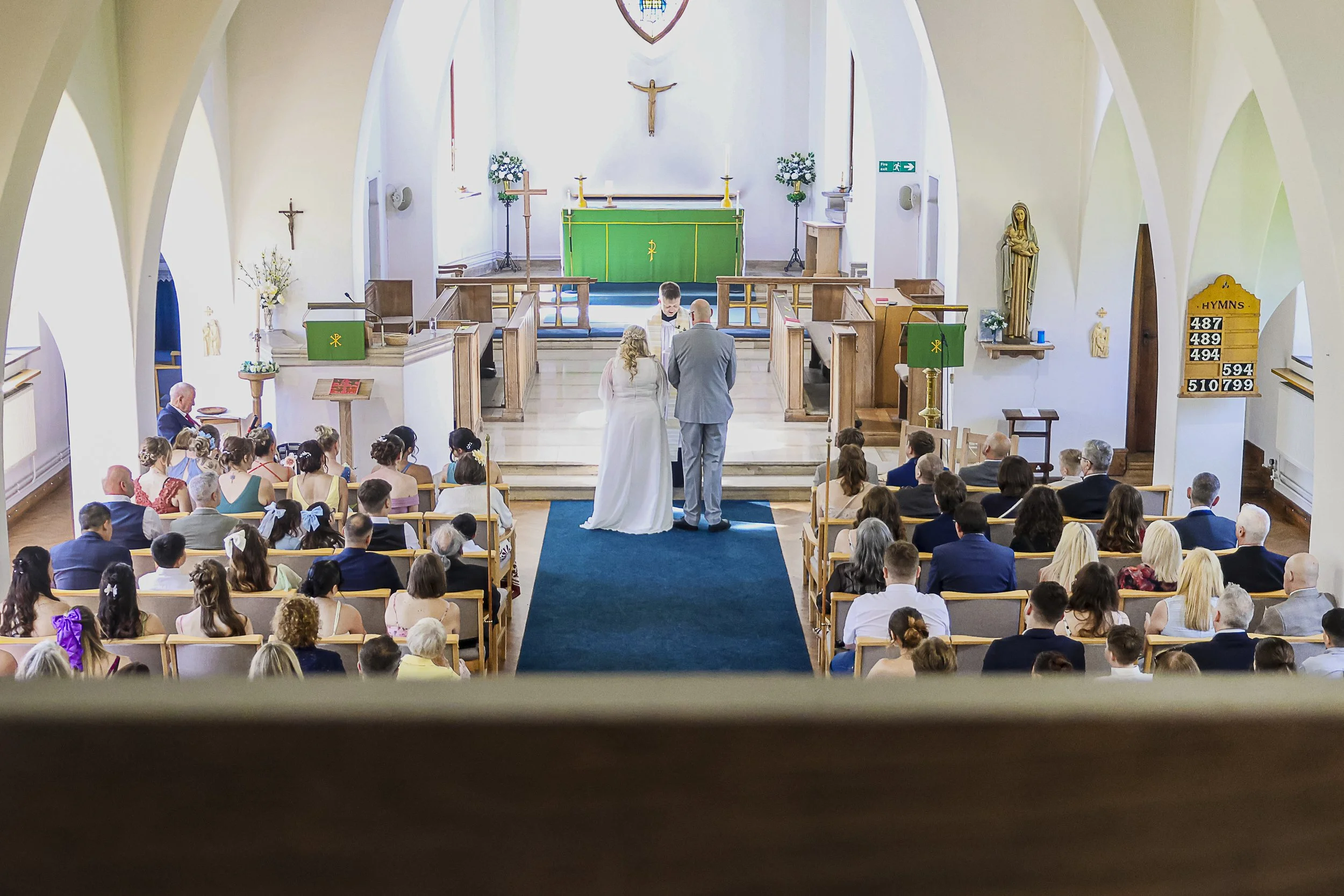 A wedding ceremony taking place inside a church with a bride and groom facing the officiant at the altar. The church is filled with guests seated in pews, and the altar features religious symbols including a crucifix and statues.