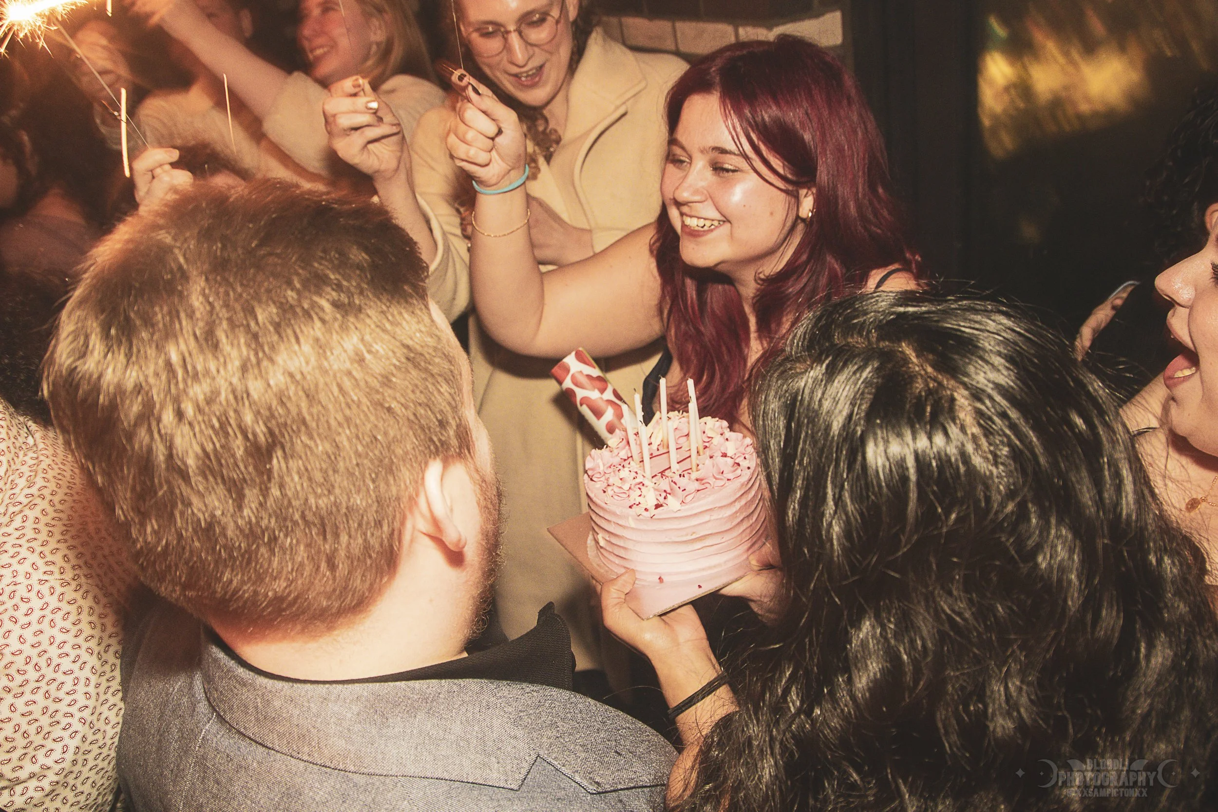 A group of people celebrating a birthday with a pink cake with candles, smiling, holding sparklers, and enjoying the party.