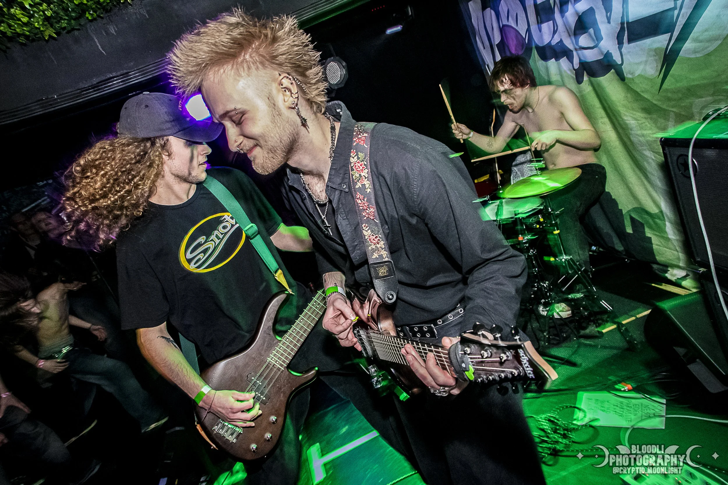 Two musicians playing electric guitars close together on stage, with a drummer in the background, under green stage lighting.