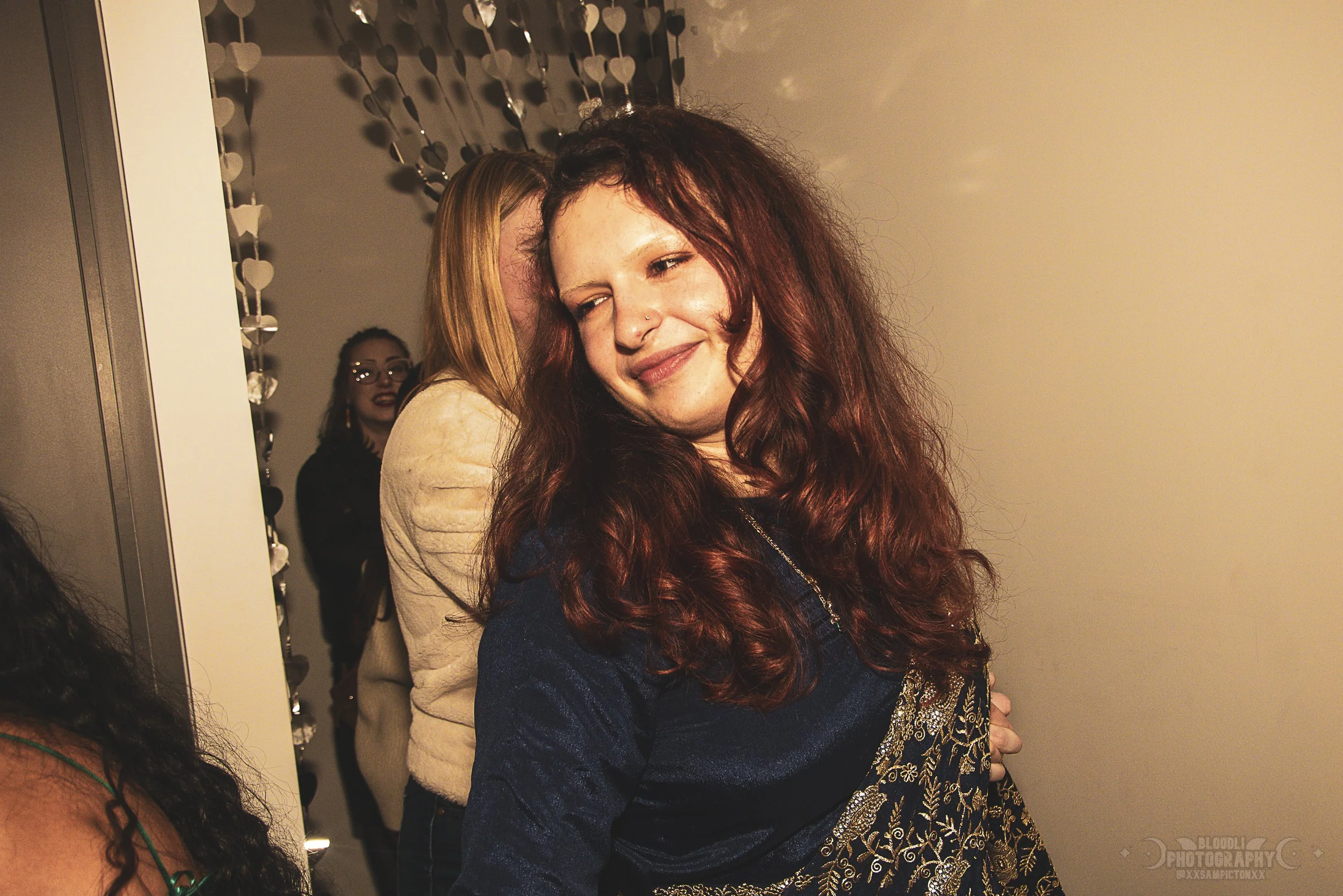 A woman with long, curly red hair smiling with her eyes closed, wearing a dark blue top with gold embroidery, at a social event with other women in the background.