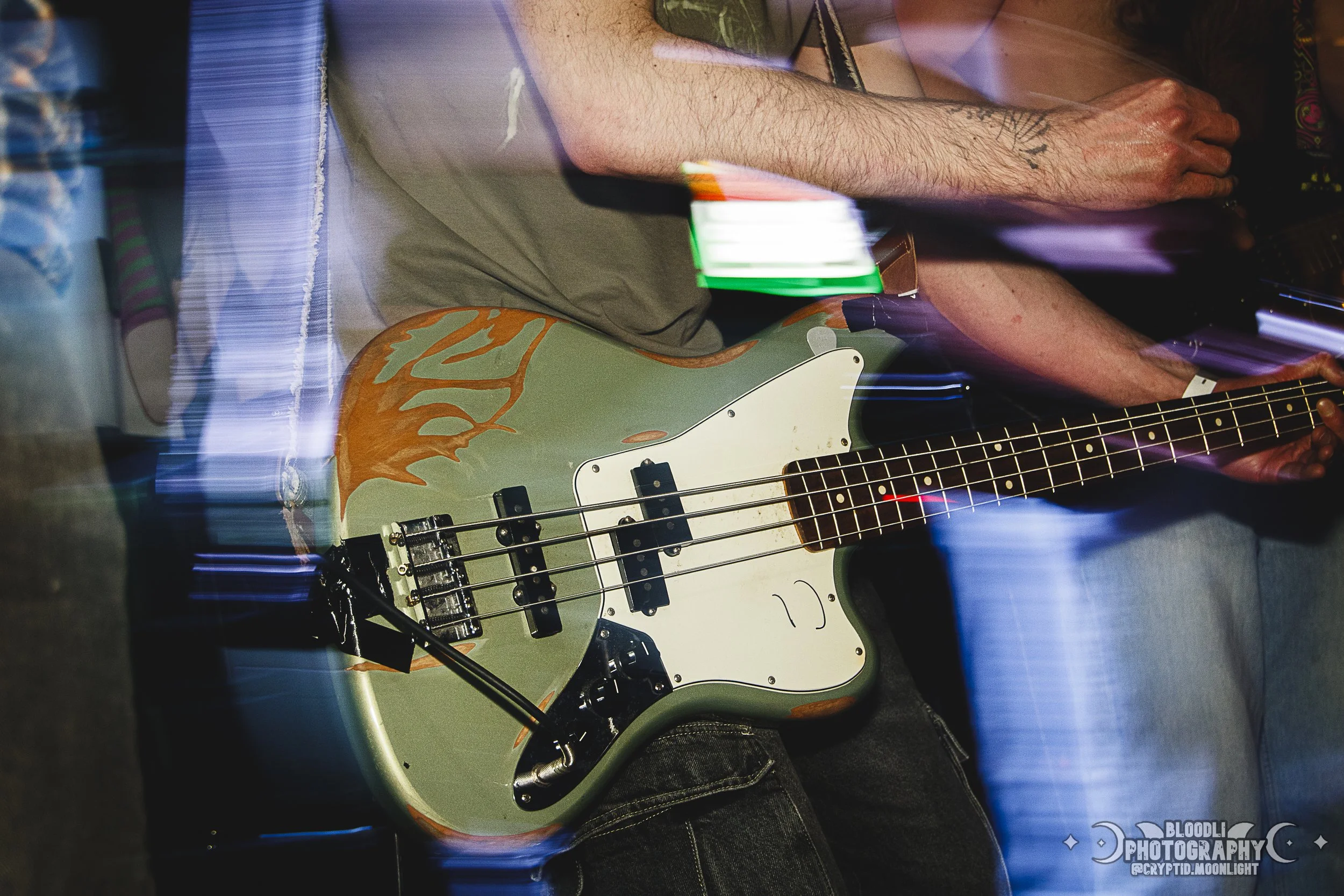 Close-up of a person's torso holding a bass guitar with a custom paint job, with colorful lighting effects.