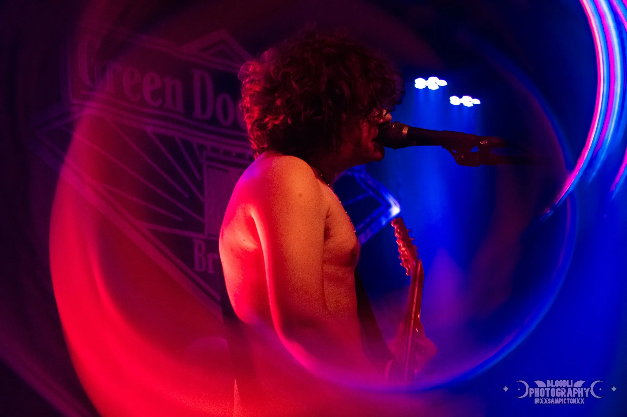 A shirtless musician has curly hair, is singing into a microphone, and playing an electric guitar on stage with colorful neon lighting and a partial logo in the background.