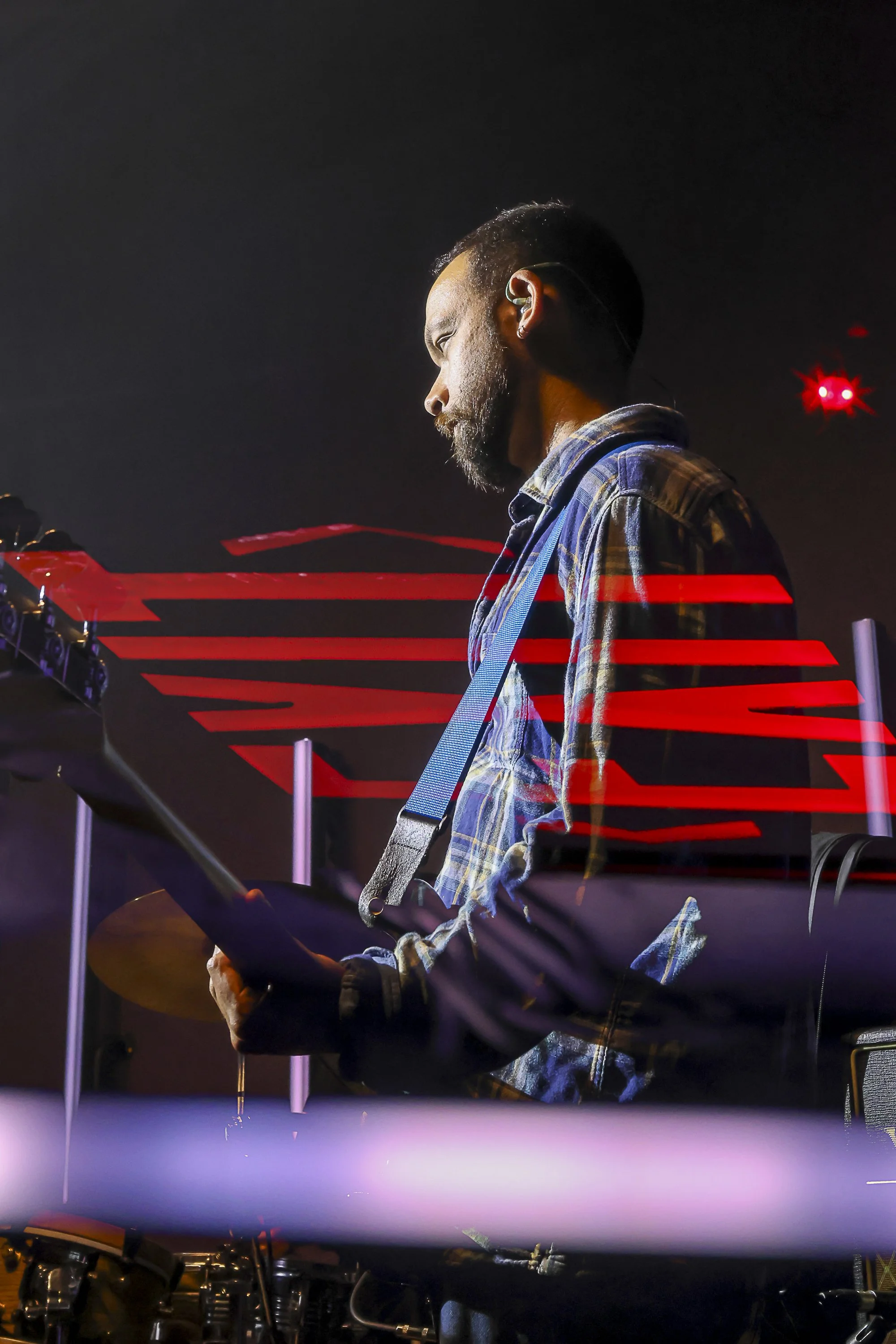 A man wearing a plaid shirt playing a guitar at night with a dark background and red light accents.