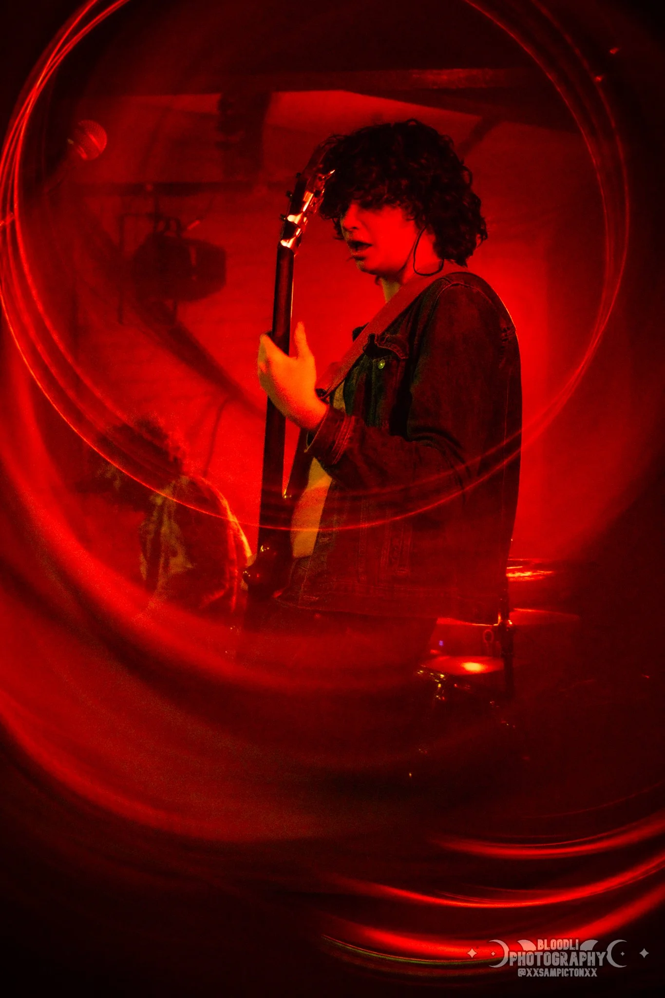 Person playing electric guitar in a red lighting setting, seen through a circular reflective surface.