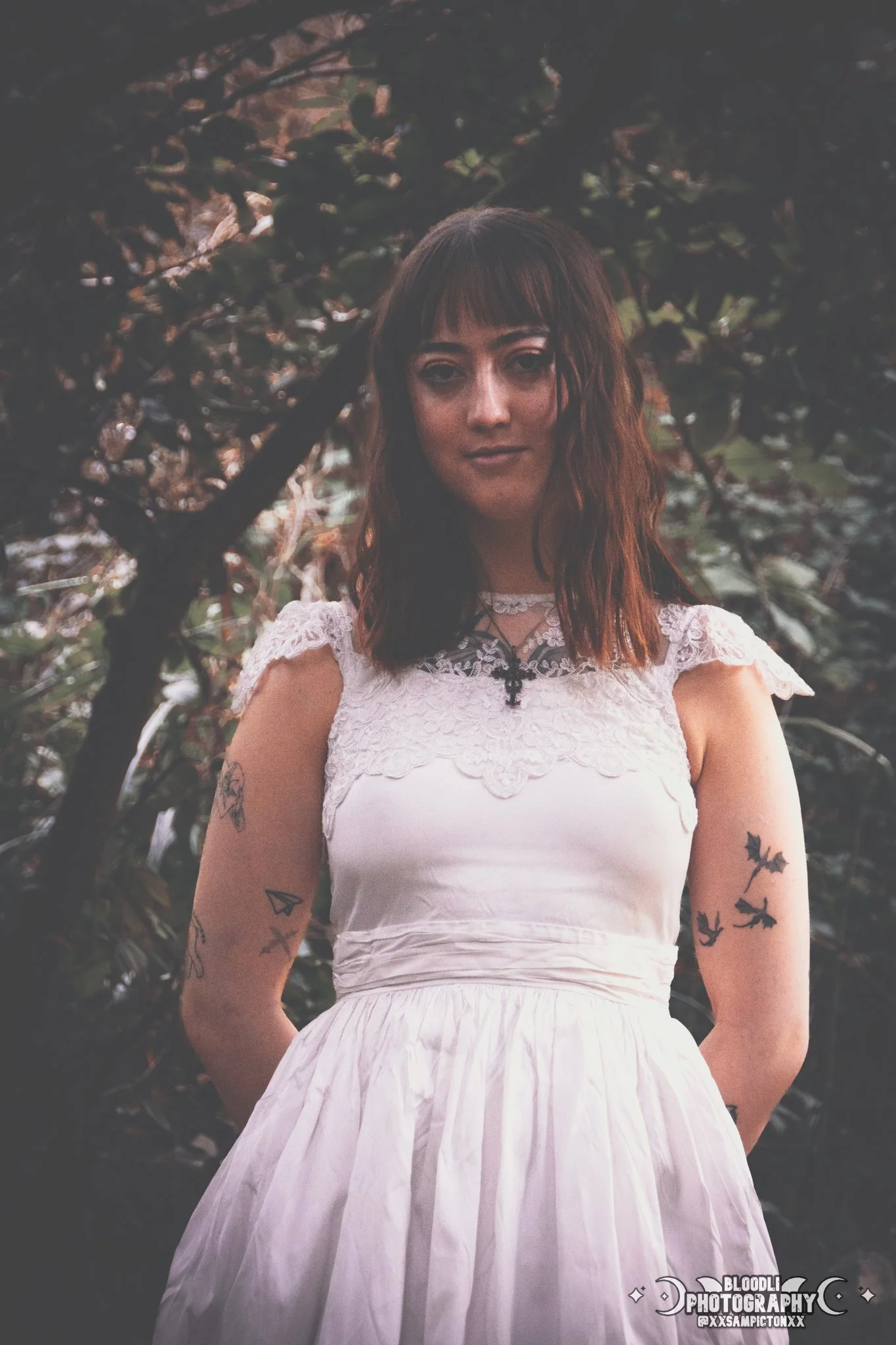 A young woman with tattoos on her arms, wearing a white lace-collared dress, stands outdoors with dark leafy trees in the background.