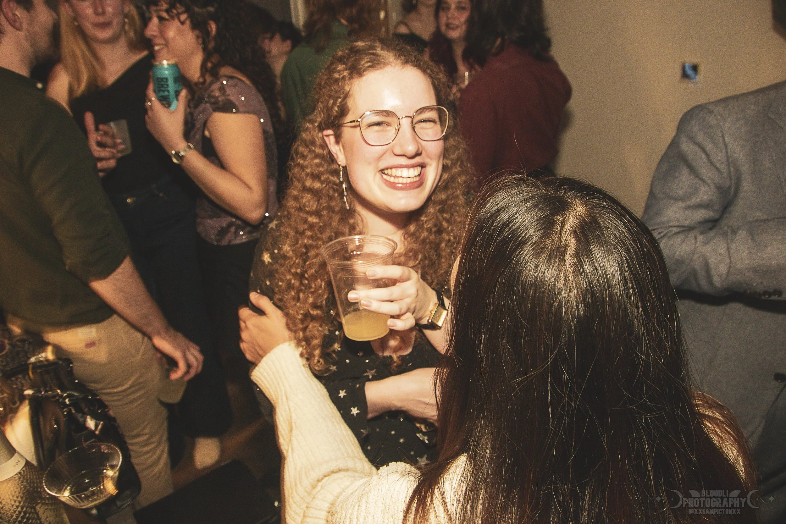 A woman with glasses and curly red hair smiling and holding a plastic cup at a party or social gathering with other people in the background.