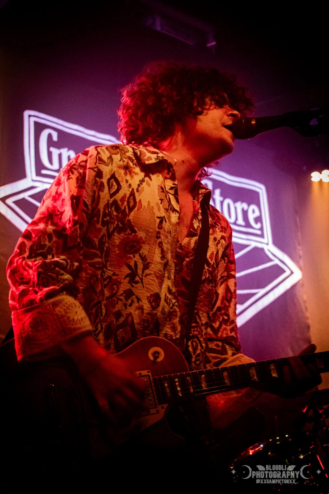 A male musician with curly hair singing into a microphone while playing an electric guitar on stage, with a neon sign in the background.