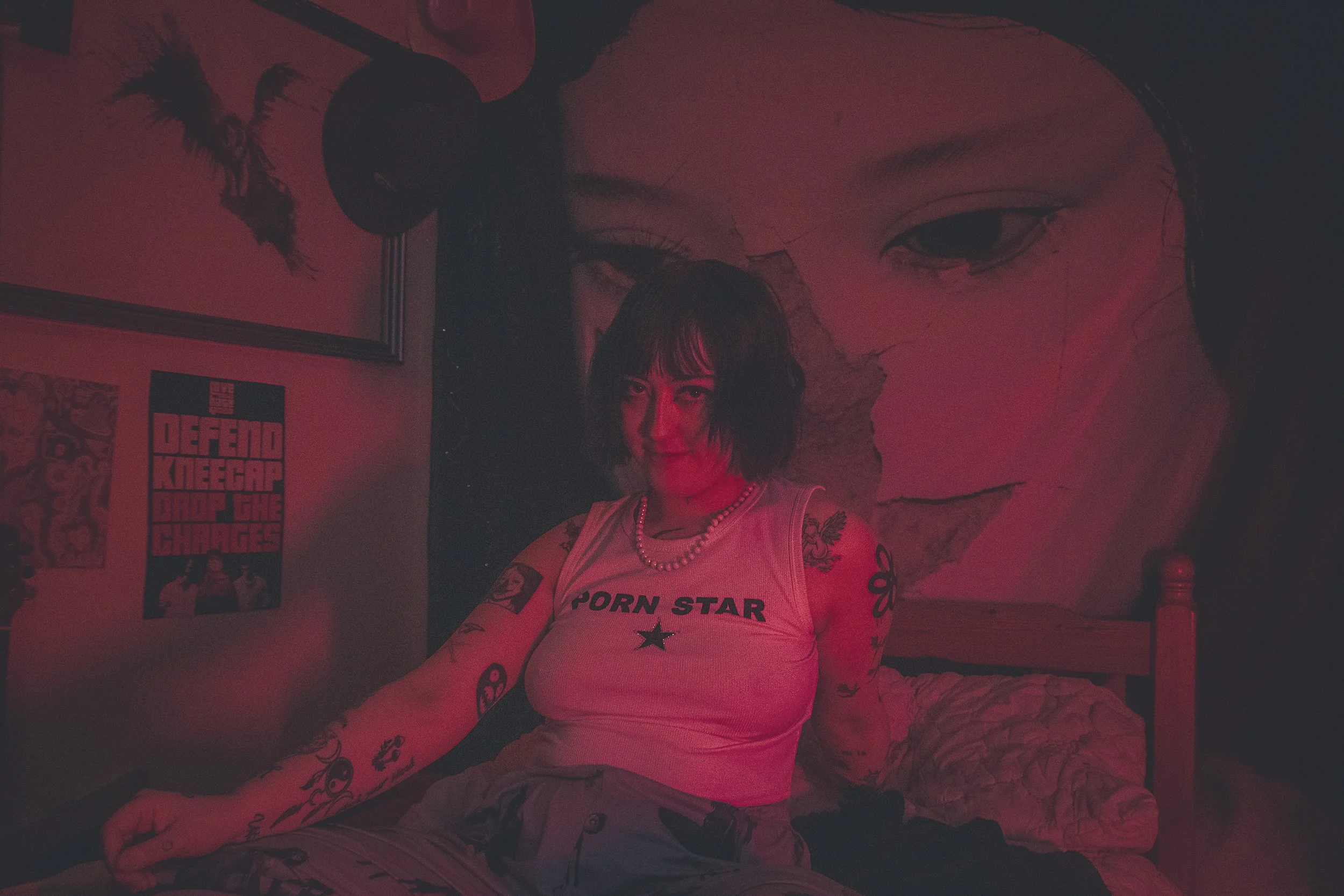 A woman with tattoos and a pearl necklace sitting on a bed in a dimly lit room with red lighting; a large mural of a woman's face with cracked paint is visible on the wall behind her.