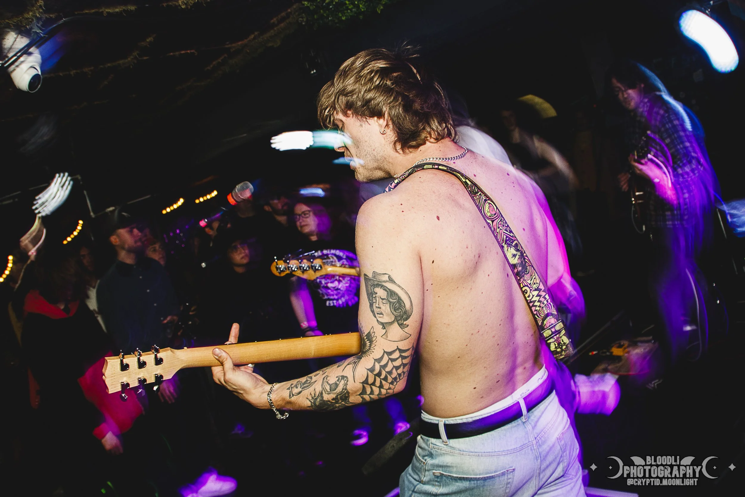 A shirtless male guitarist with tattoos playing guitar at a concert, audience watching in the background, with purple and blue stage lighting.