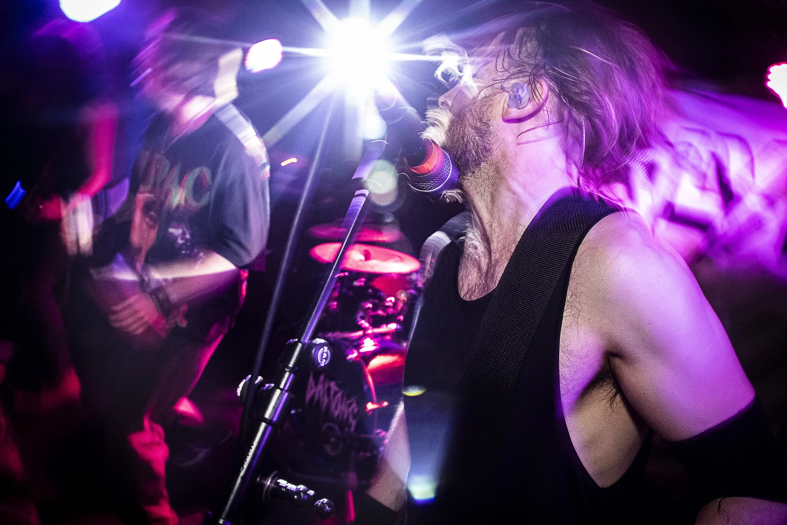 A rock band performing on stage with vibrant purple and pink lighting, featuring a vocalist with long hair and a sleeveless shirt singing into a microphone.
