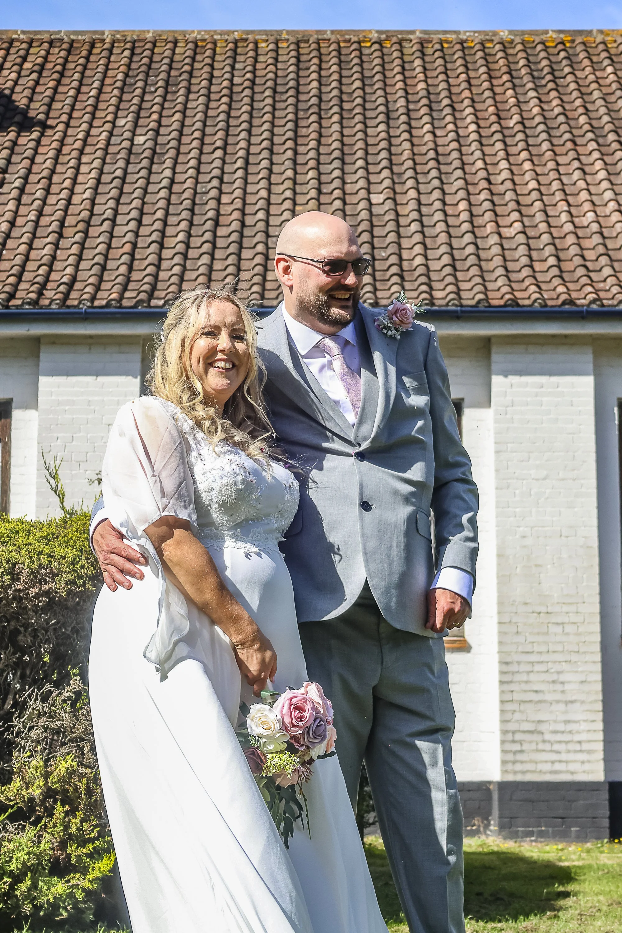A happy couple, a woman in a white wedding dress holding a bouquet of pink and white roses and a man in a gray suit with a pink tie and boutonniere, standing outdoors in front of a white brick building with a tiled roof under a clear blue sky.