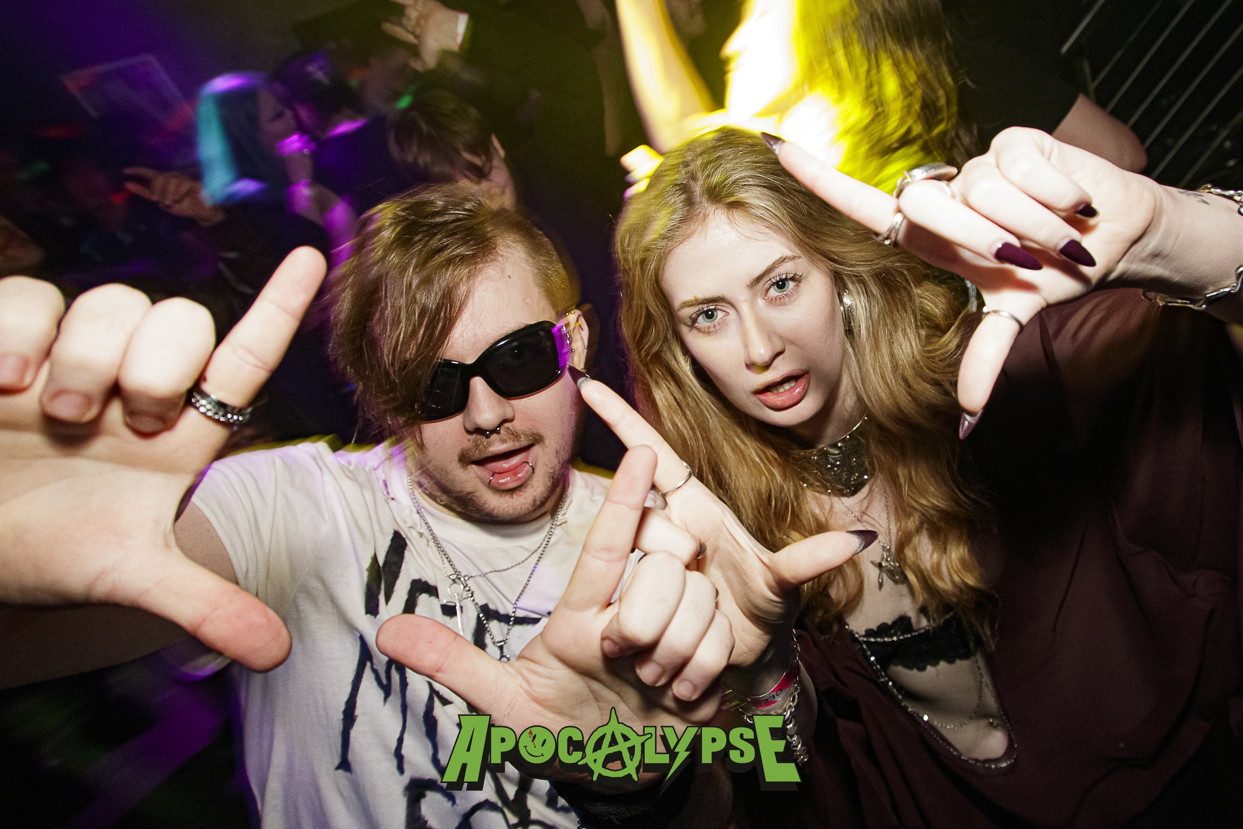 Two people at a party or concert, making hand gestures and looking at the camera. The person on the left has dark sunglasses, a white T-shirt, and dark hair. The person on the right has long, wavy blonde hair and is wearing a dark top. The background