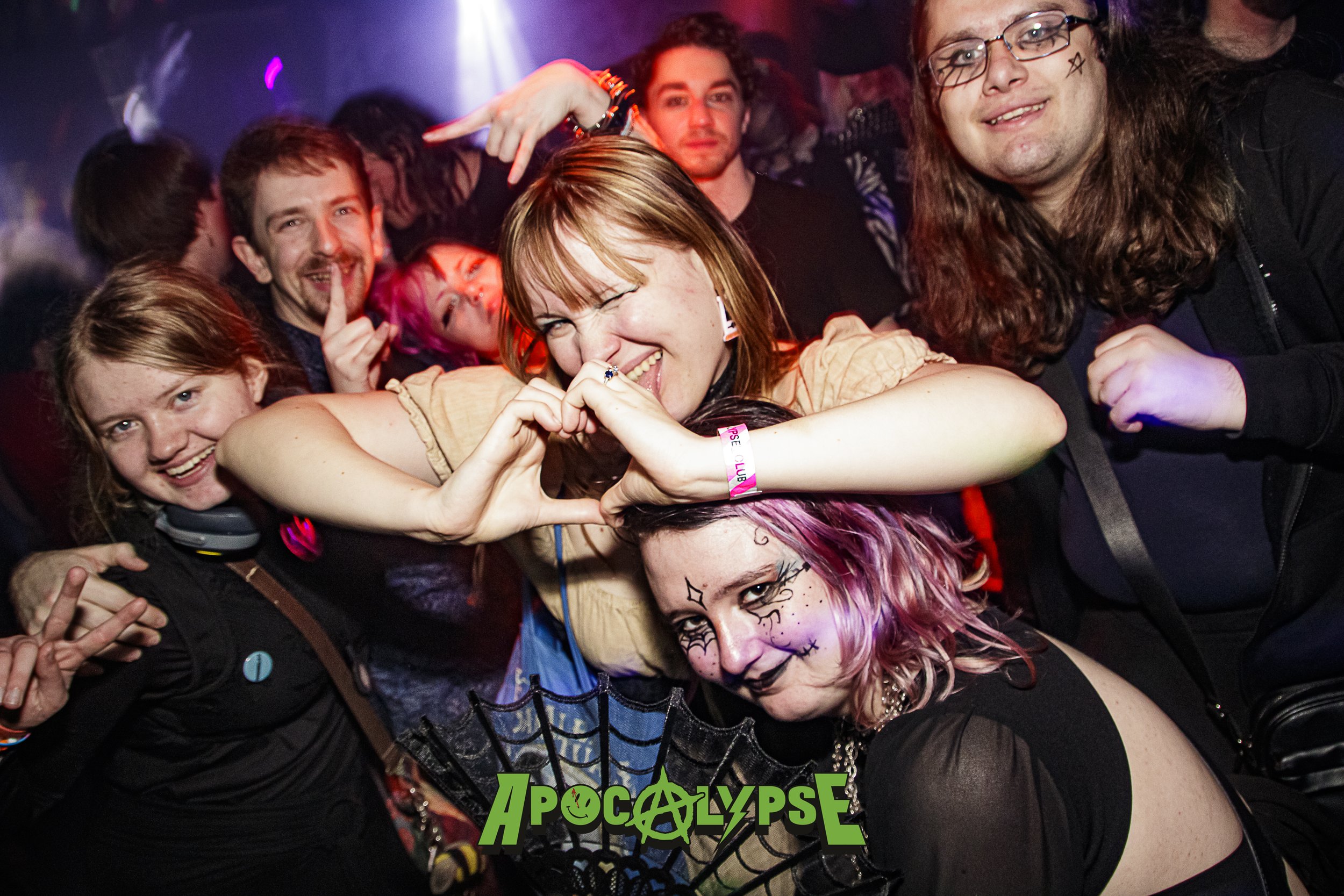 A group of people at a party or concert, with some making playful and expressive faces. The woman in the center is making a heart shape with her hands, and the woman in front has face makeup with a gothic or Halloween theme. The crowd appears happy a