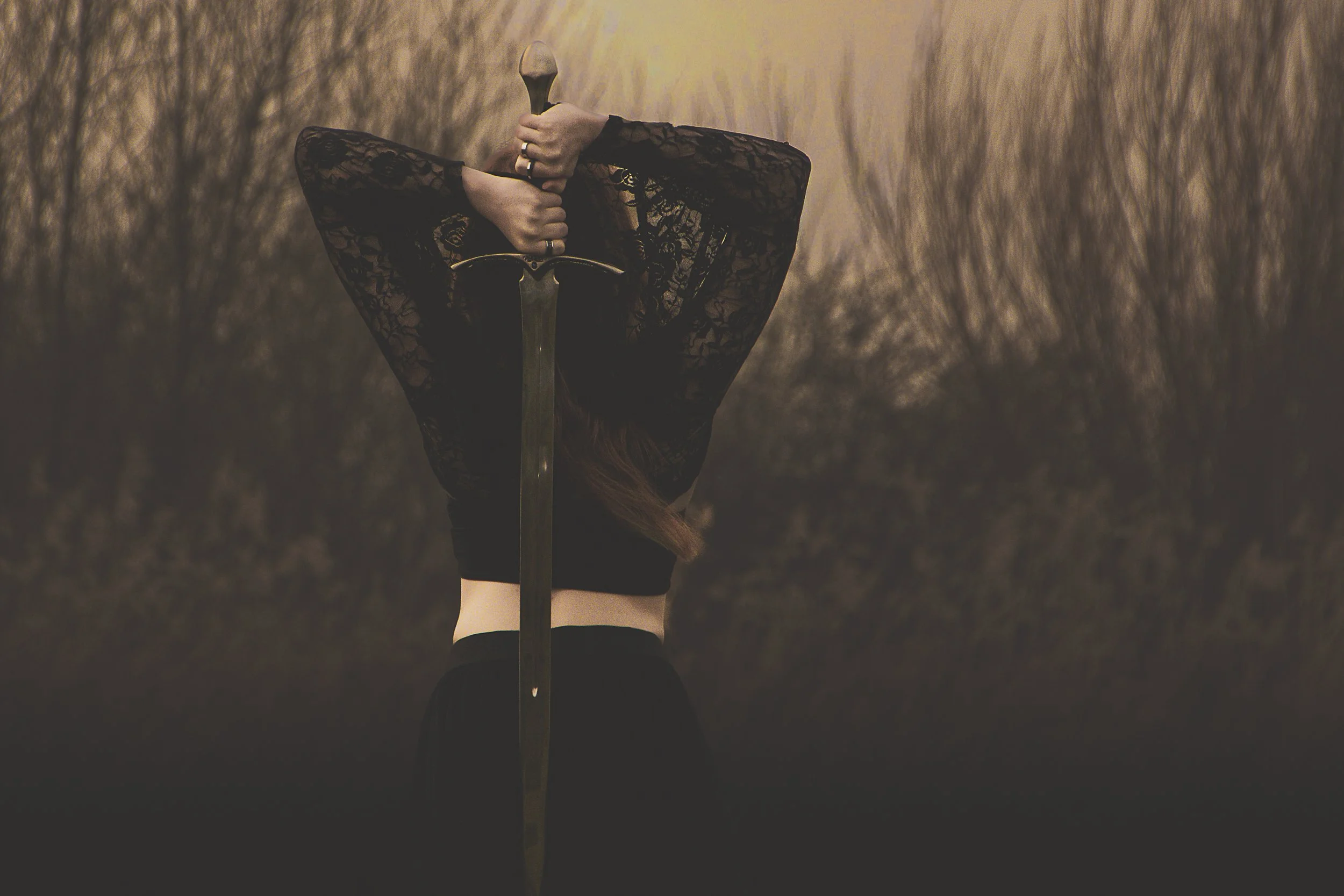 Person in black lace top holding a sword upright in front of a blurred wooded background