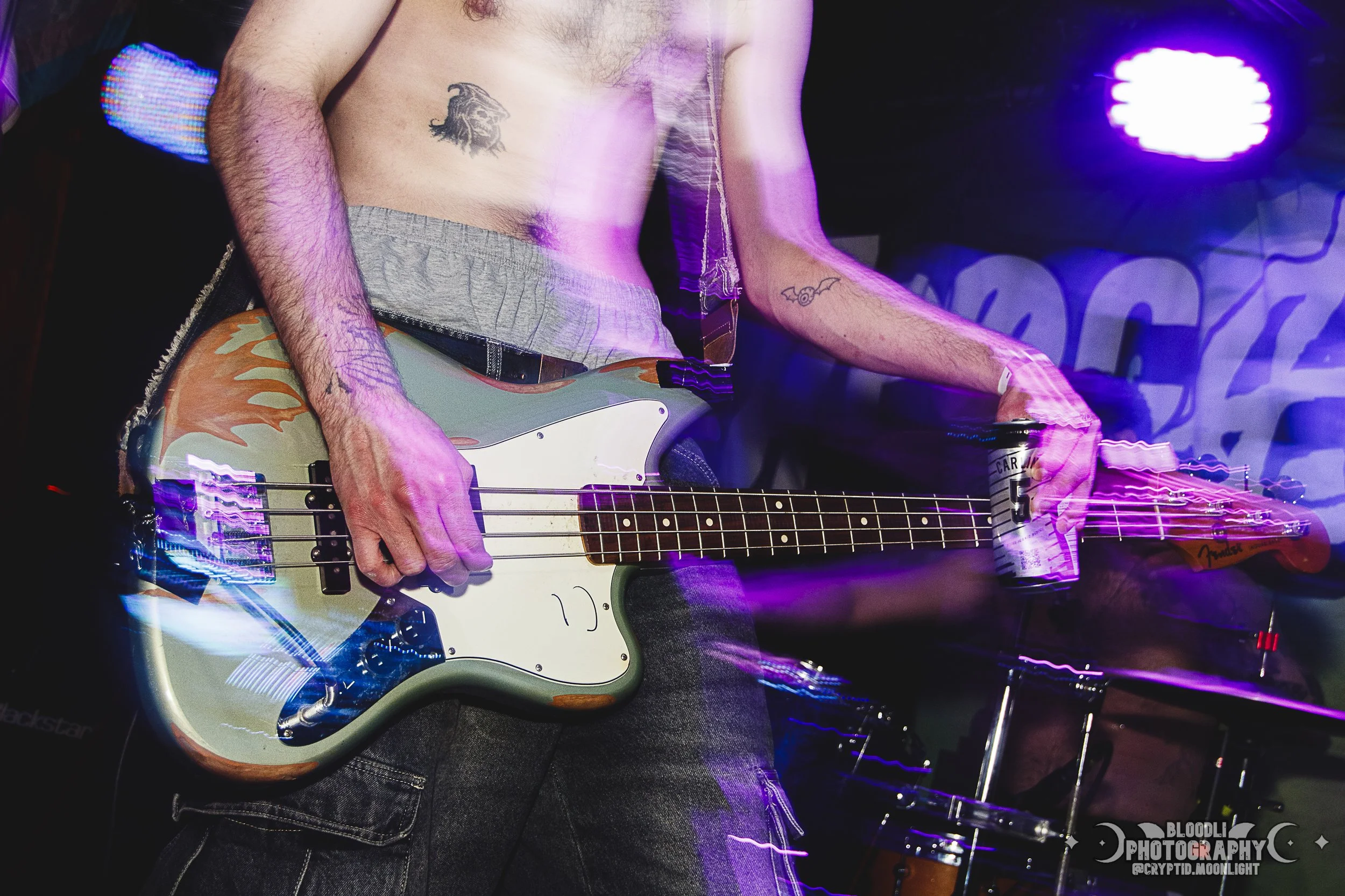 A shirtless man playing a Fender bass guitar during a live music performance, with purple and pink stage lights creating a blurred motion effect.