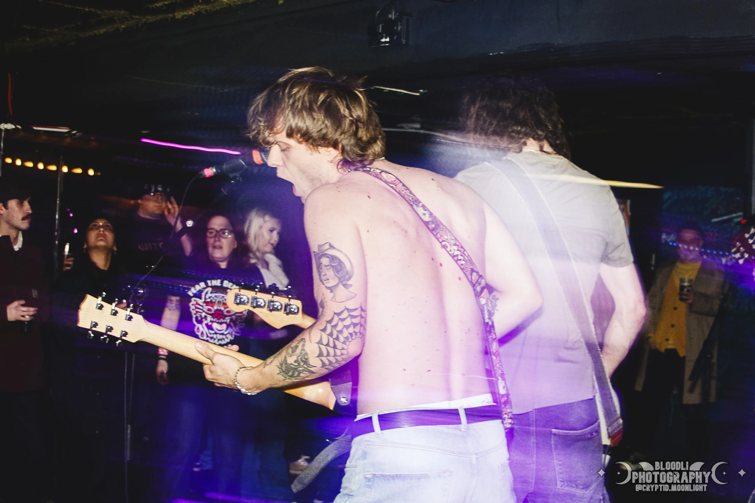 A shirtless male guitarist performing on stage with audience members watching in the background, colorful lighting, and motion blur effects.