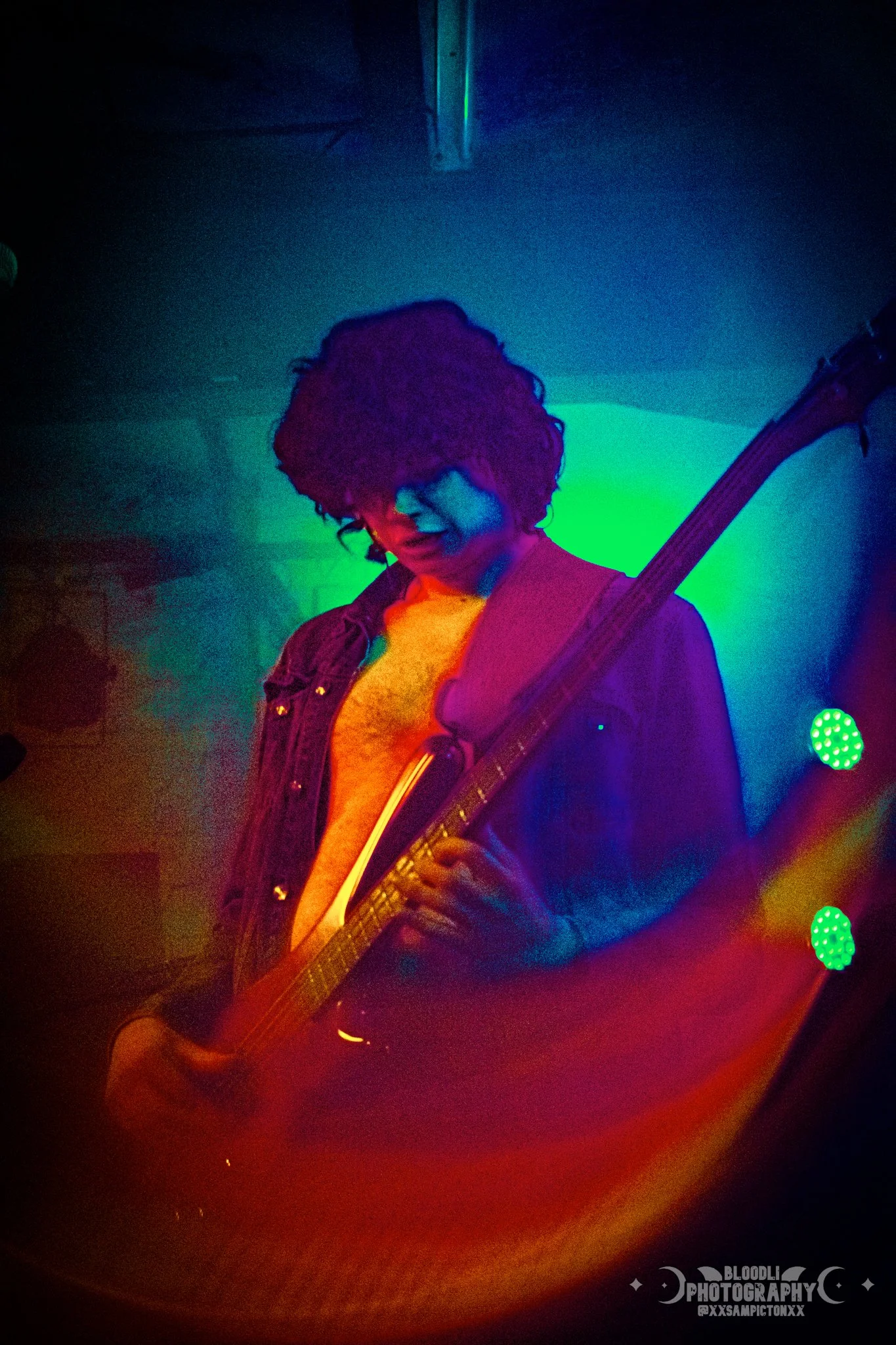 A person playing an electric guitar during a live music performance. The scene is illuminated with vibrant, colorful stage lights including green, blue, purple, and red, creating a moody and lively atmosphere.