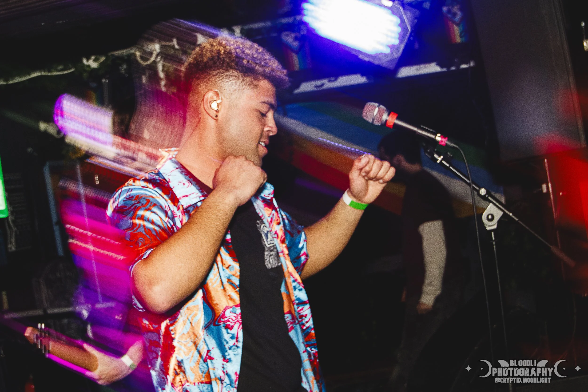 Young man dancing with closed eyes at a nightclub, colorful lights and a microphone stand nearby.