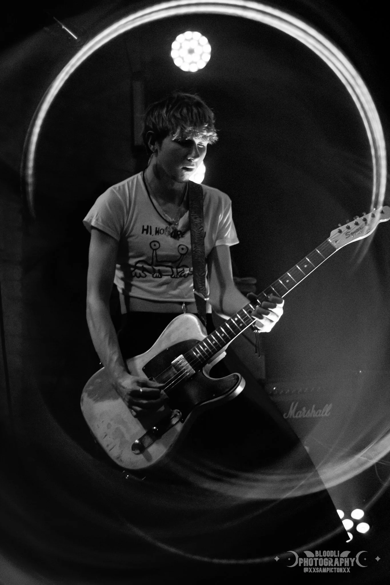 A black and white photo of a person playing an electric guitar on stage, with a Marshall amplifier in the background, illuminated by stage lights.