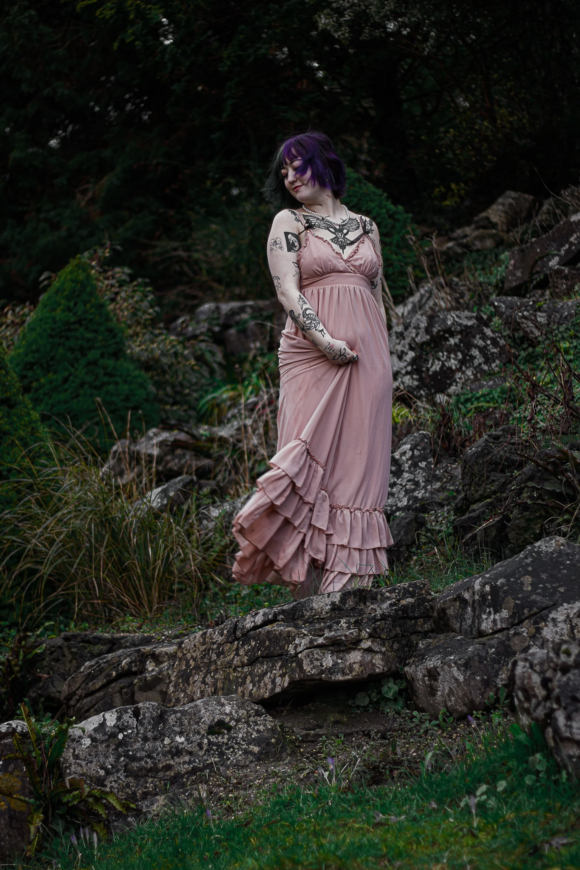 A woman with purple and black hair, wearing a pink dress with ruffles, stands outdoors on rocky terrain surrounded by green foliage and trees.