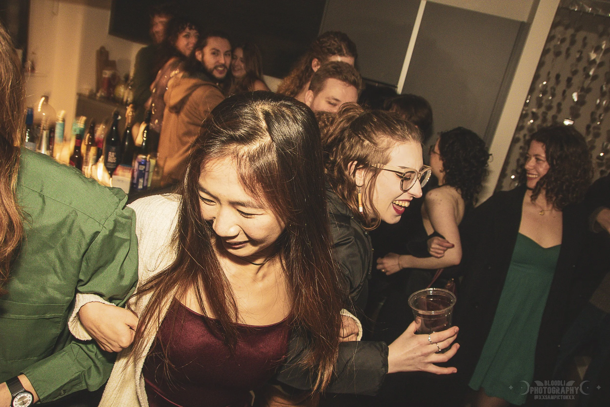 Group of people at a party, some dancing and holding drinks, with a bar in the background.