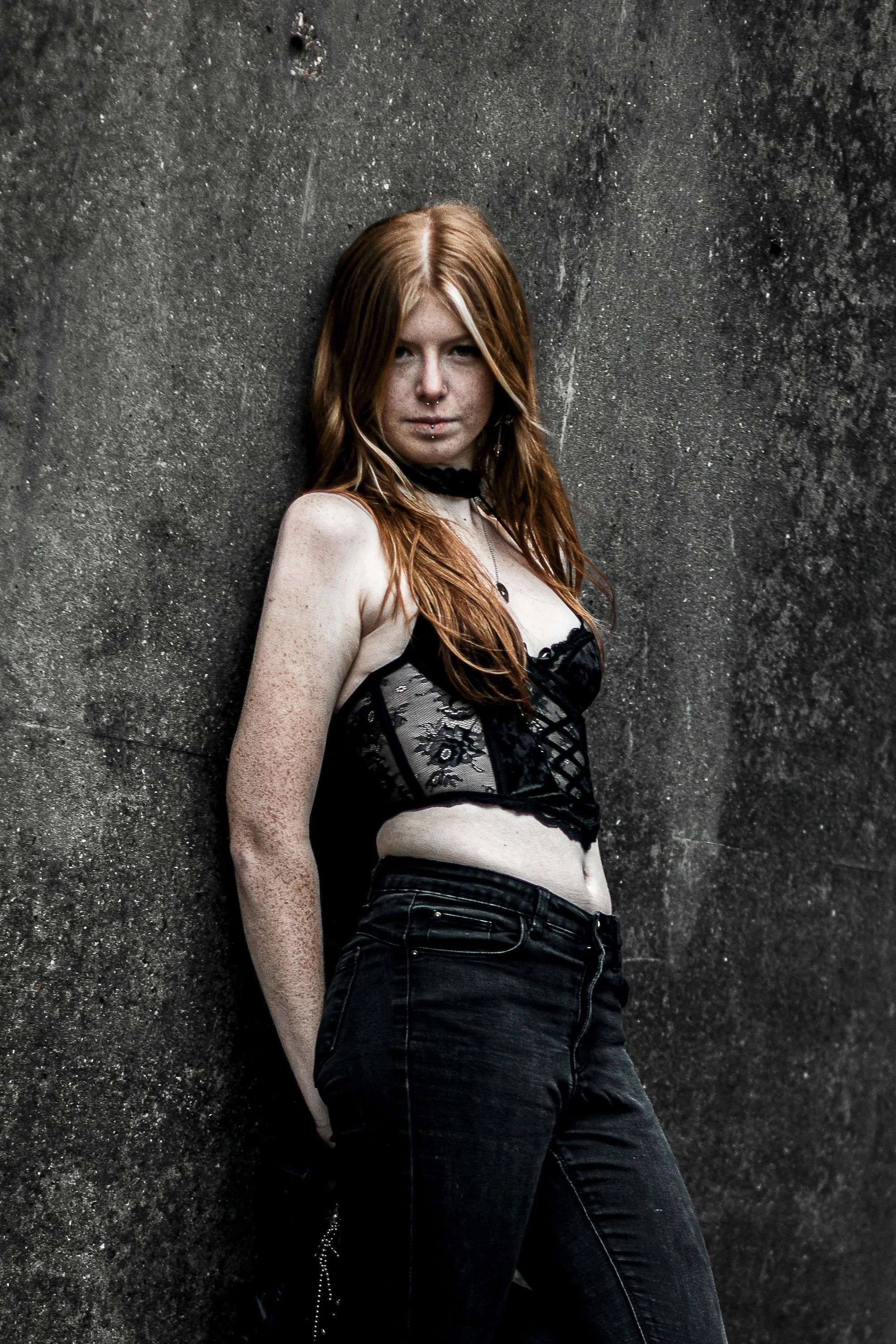 A young woman with red hair, freckles, and piercings, wearing a black lace bustier and black jeans, leaning against a textured dark wall.