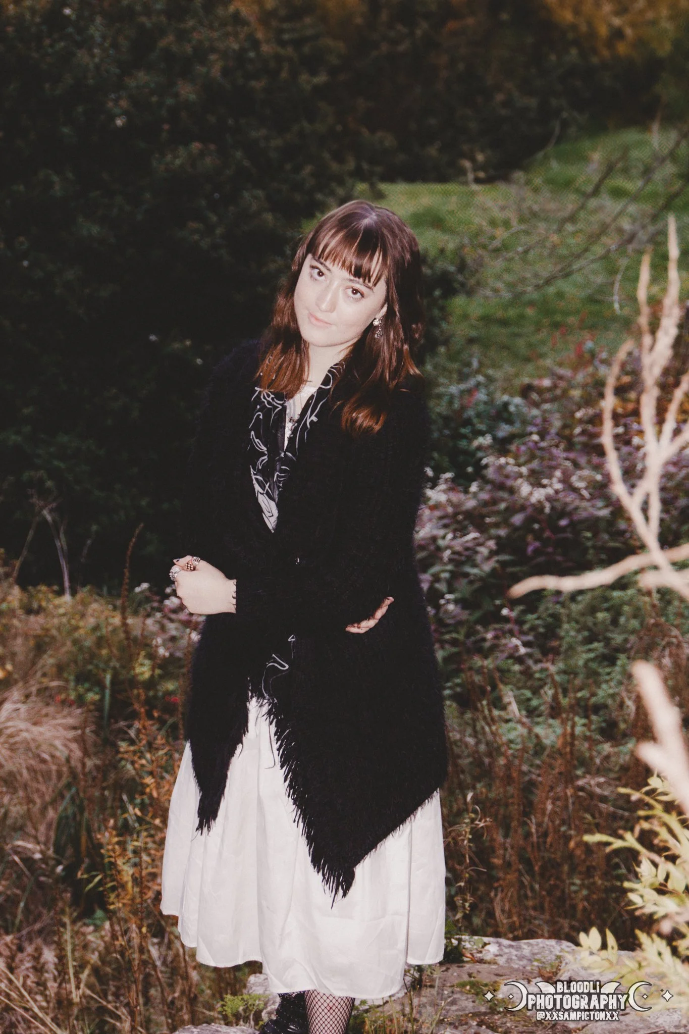 A woman with brown hair and bangs stands outdoors among trees and bushes, wearing a black coat over a white dress with fishnet stockings.