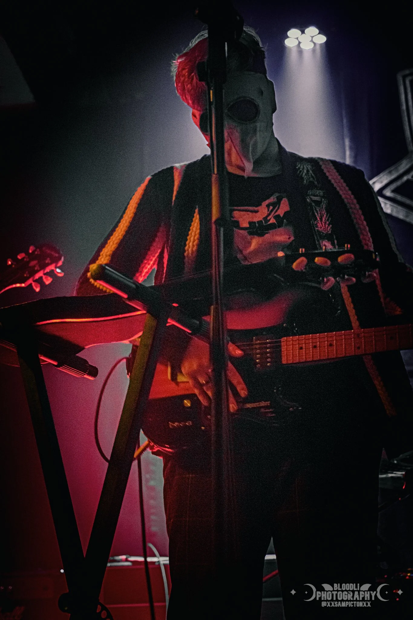 Musician on stage playing guitar, wearing a mask and a jacket with colorful stripes, illuminated by red and purple stage lights.