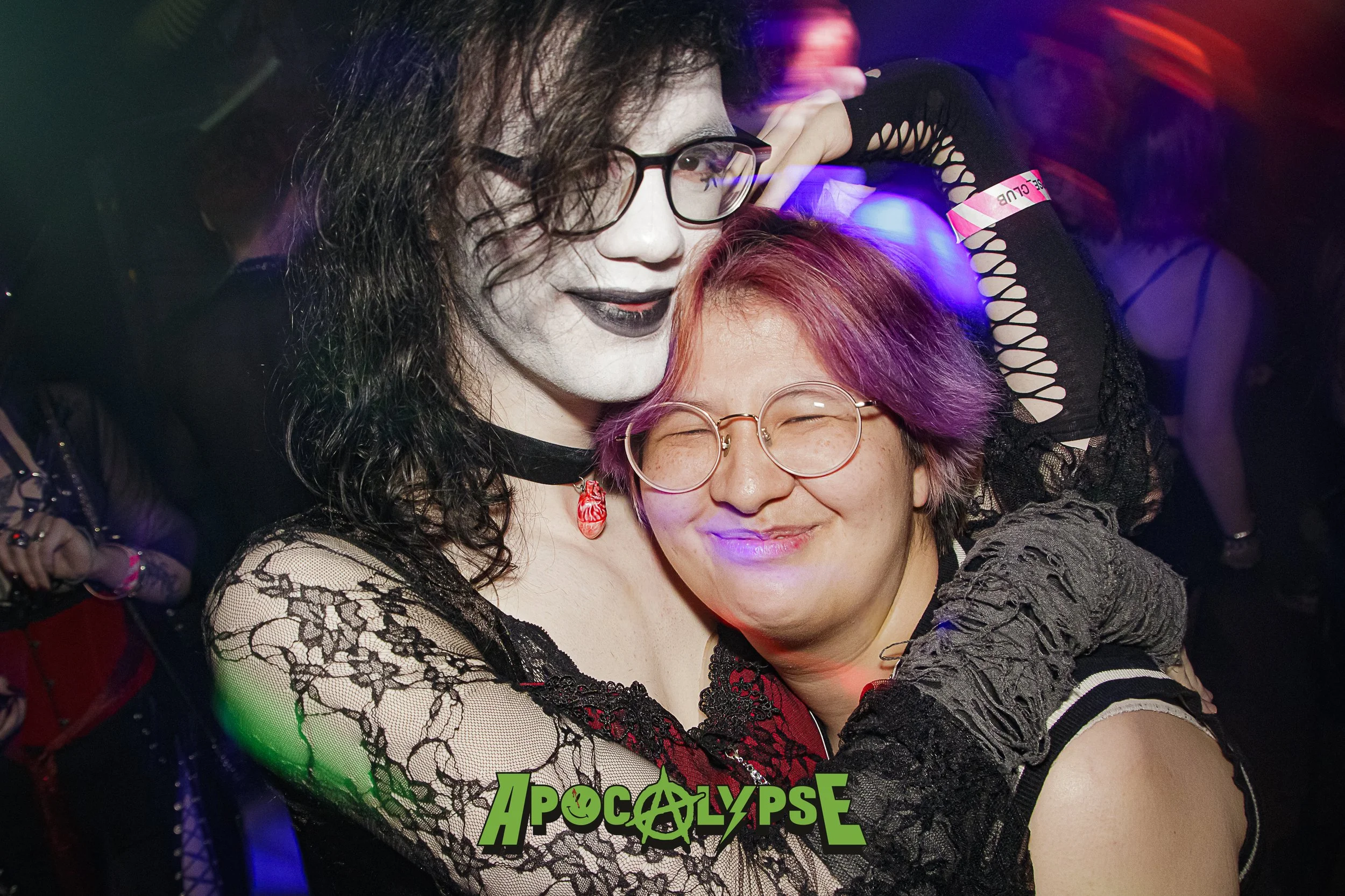 Two people dressed in costumes at a party, one with face makeup resembling a corpse, and the other with purple hair and glasses, smiling and hugging each other. The background is dark with colorful lights, and the word 'Apocalypse' is displayed at th