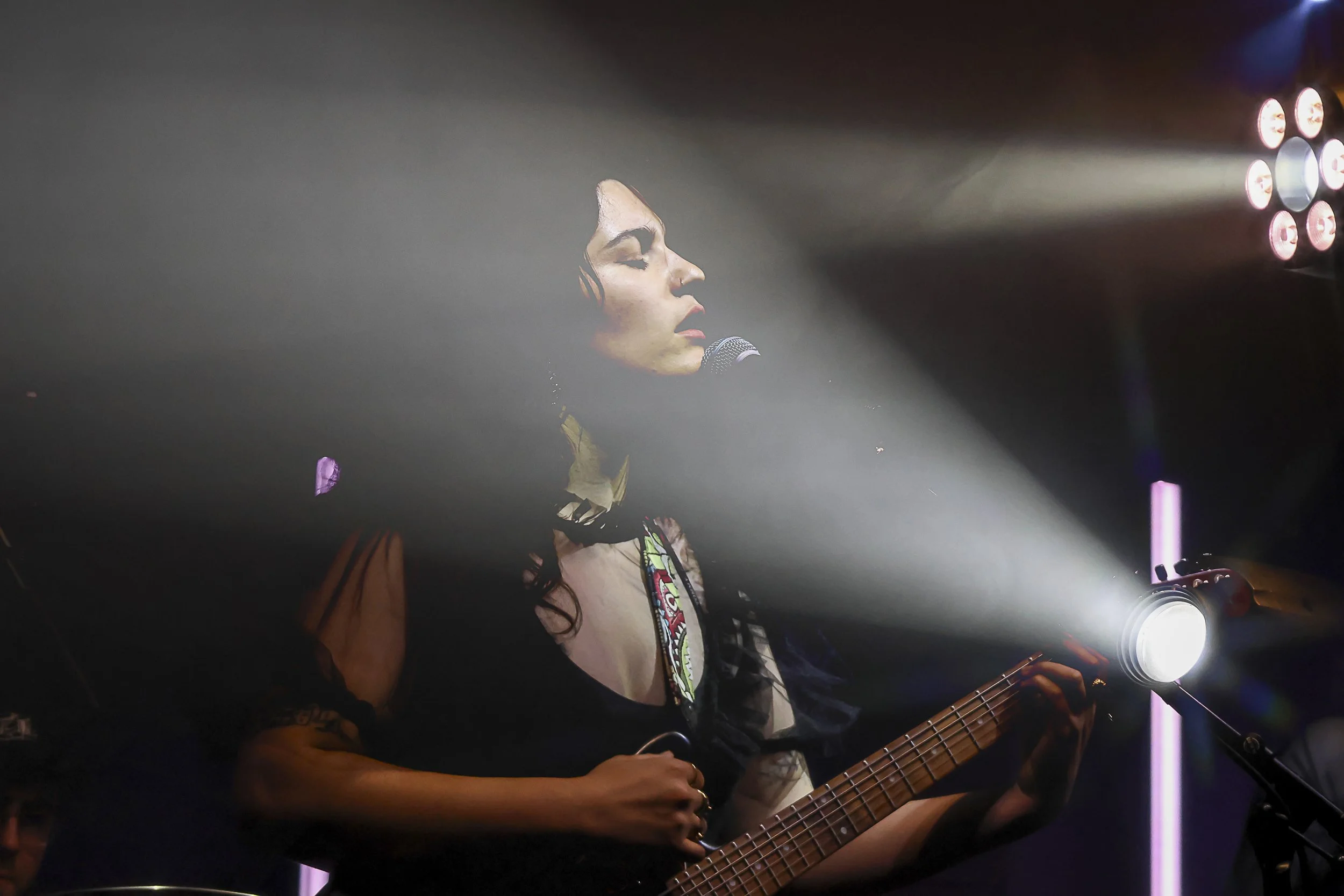 A woman performing live on stage, singing into a microphone with her eyes closed, illuminated by stage lights, while playing an acoustic guitar.