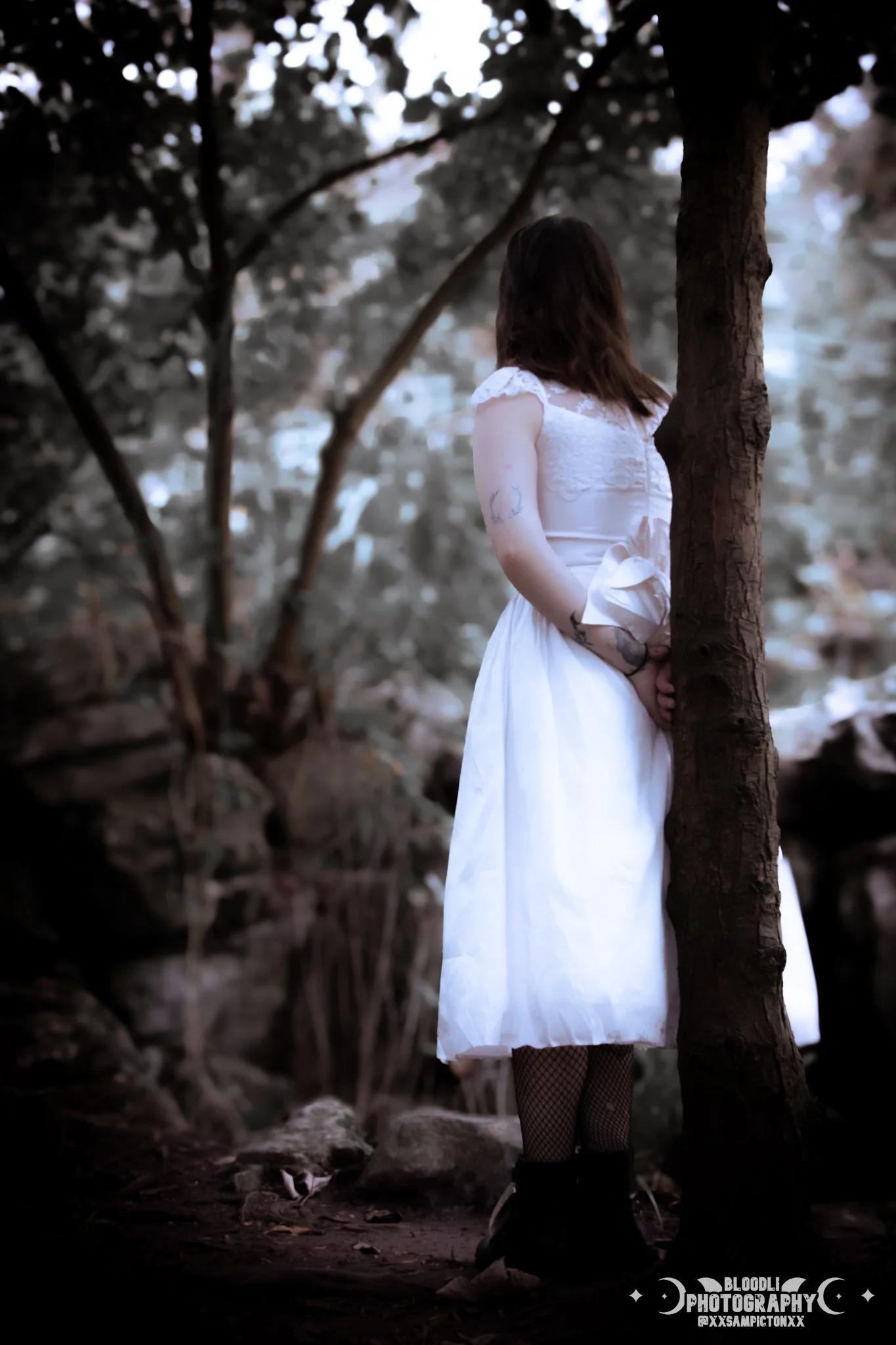 A woman in a white dress and fishnet stockings standing next to a tree outdoors, with a blurred natural background.