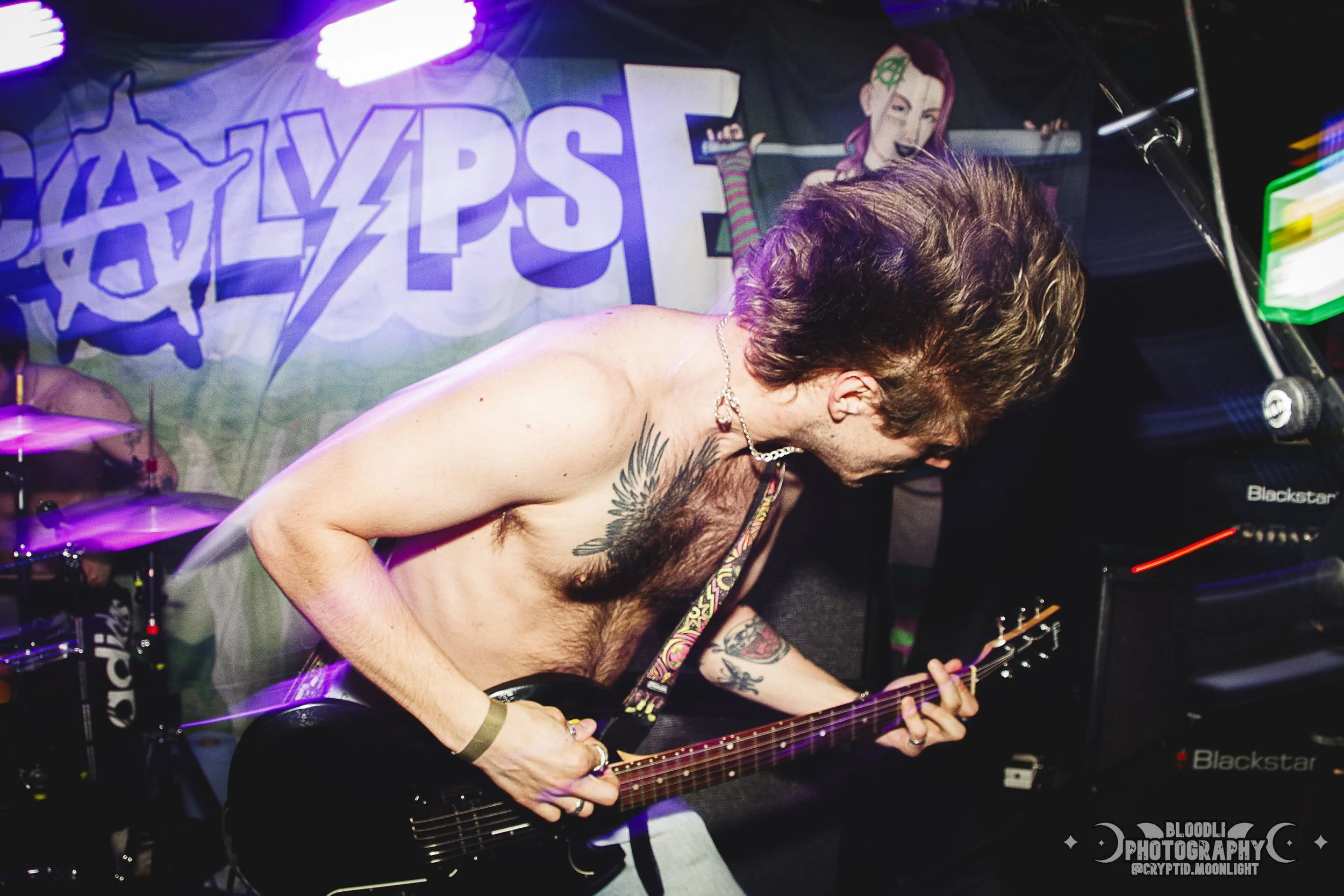 A shirtless guitarist with tattoos playing an electric guitar on stage during a live performance with a 'GAYLZZPS' band banner in the background.