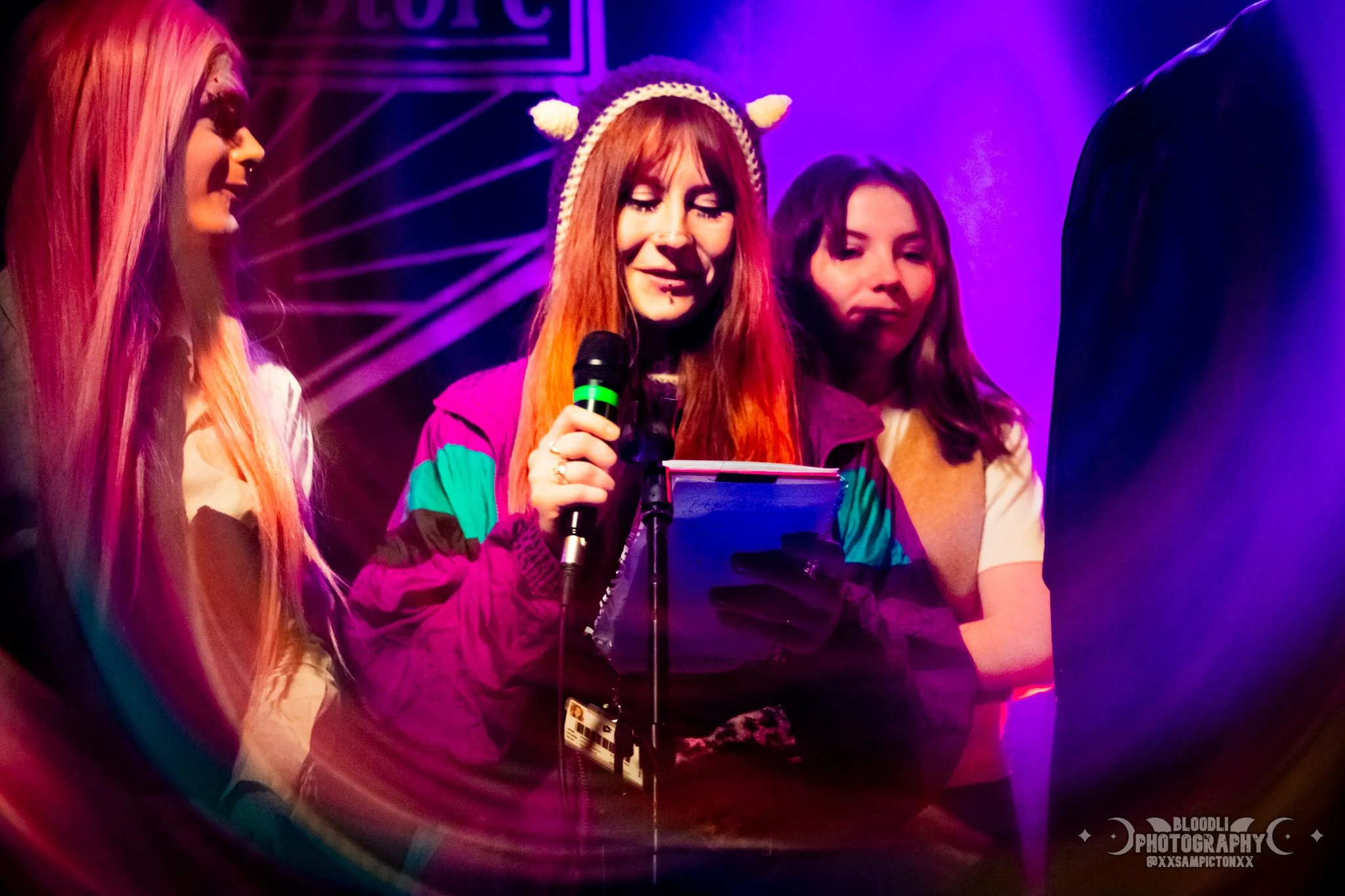 Four women on stage, with colorful lighting, one in the center holding a microphone and a notebook, wearing a headband with pom-poms and a colorful jacket, appearing to read or speak.