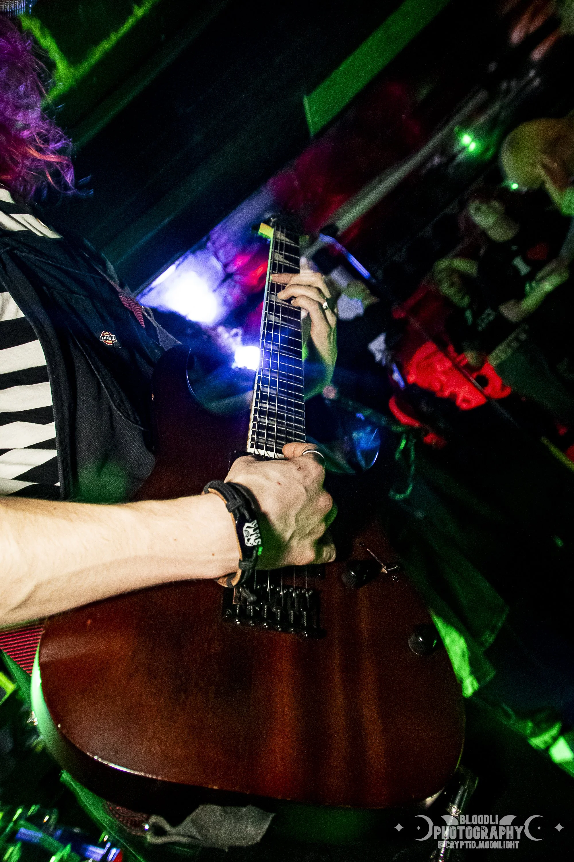 A person playing an electric guitar in a dark, colorful nightclub with green, purple, and red lights, and a small audience watching.