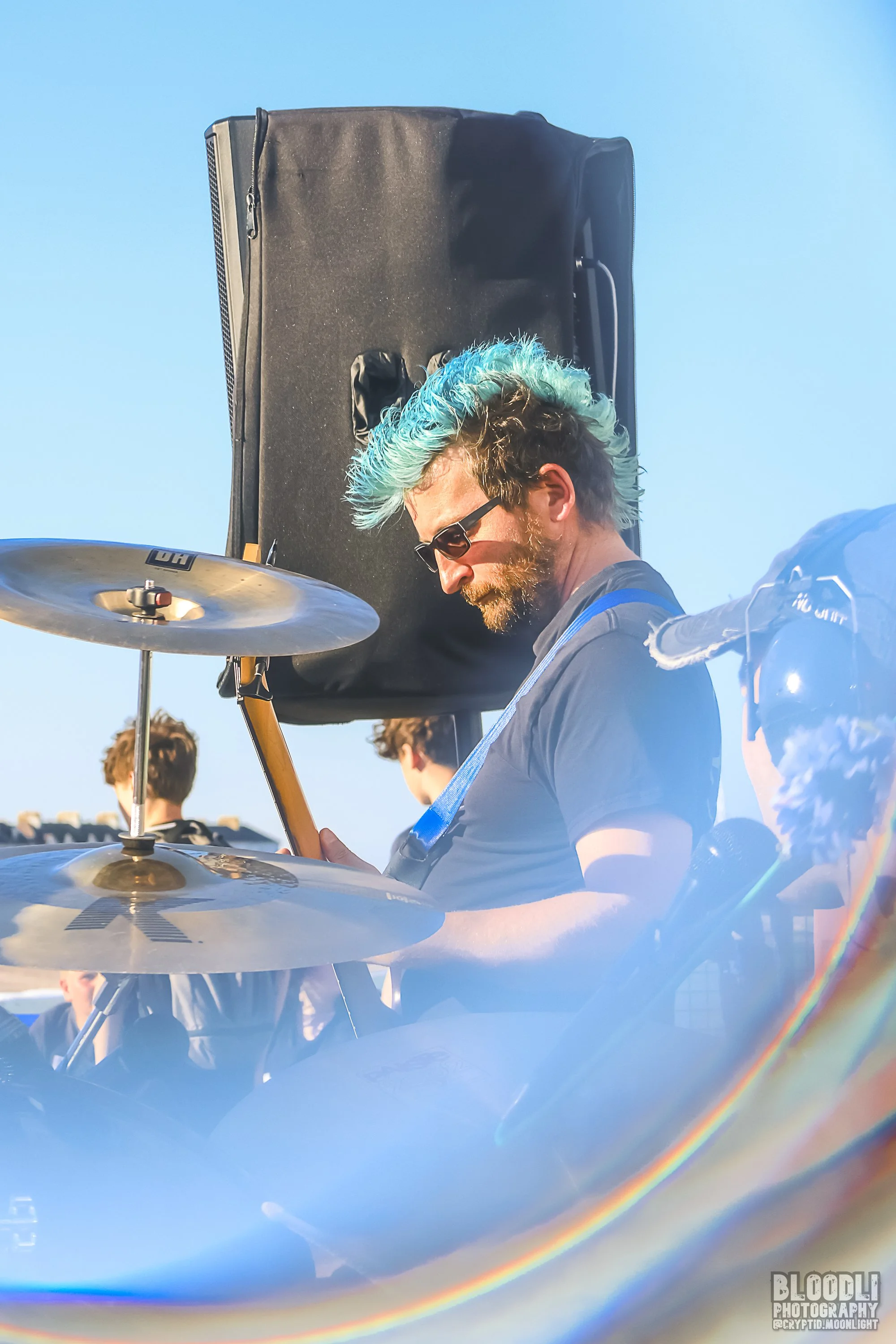 A man with colorful blue and green hair wearing sunglasses playing a drum set outdoors against a clear blue sky.
