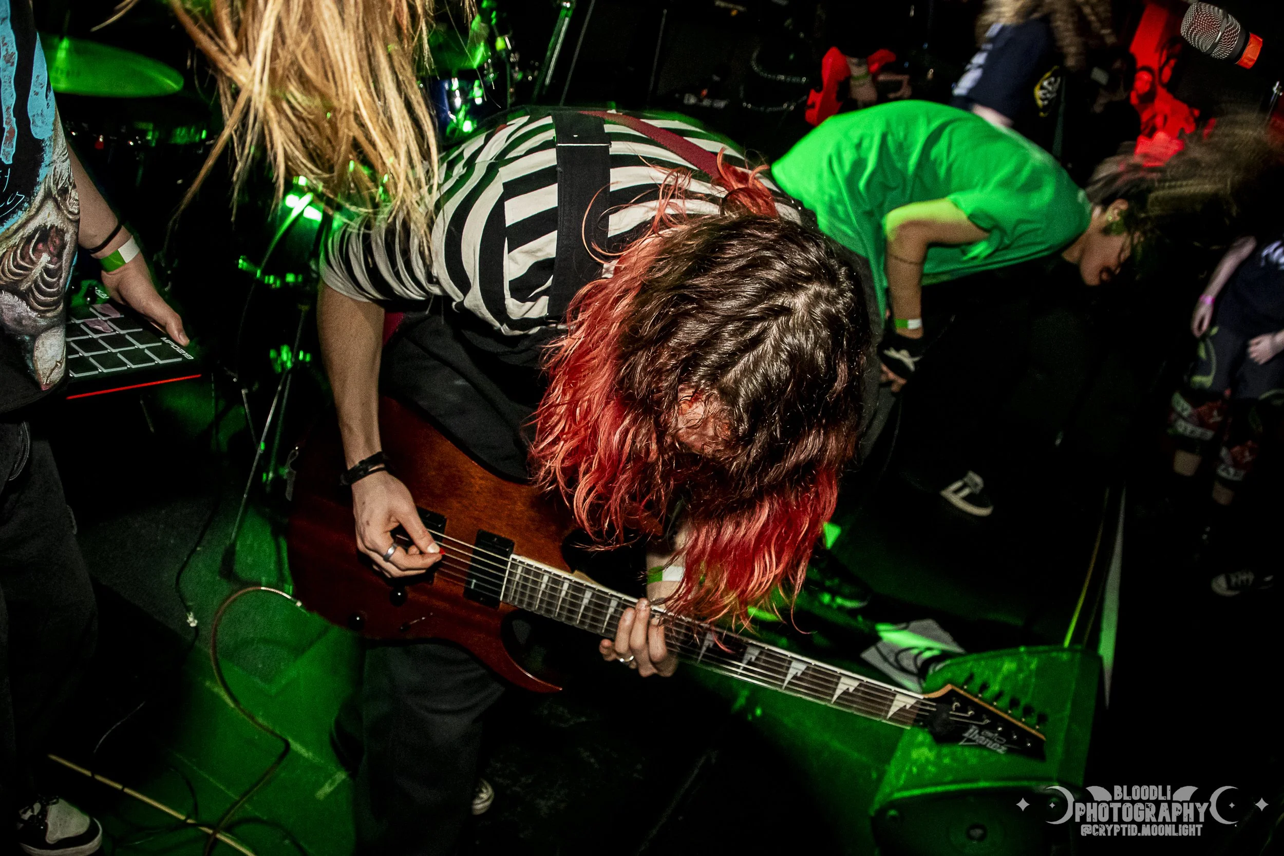 A person plays an electric guitar on stage with long, wavy hair dyed red at the tips, wearing a black and white striped shirt. There are people in the background in a dark setting with green lighting, and some are bent over or focused on equipment.