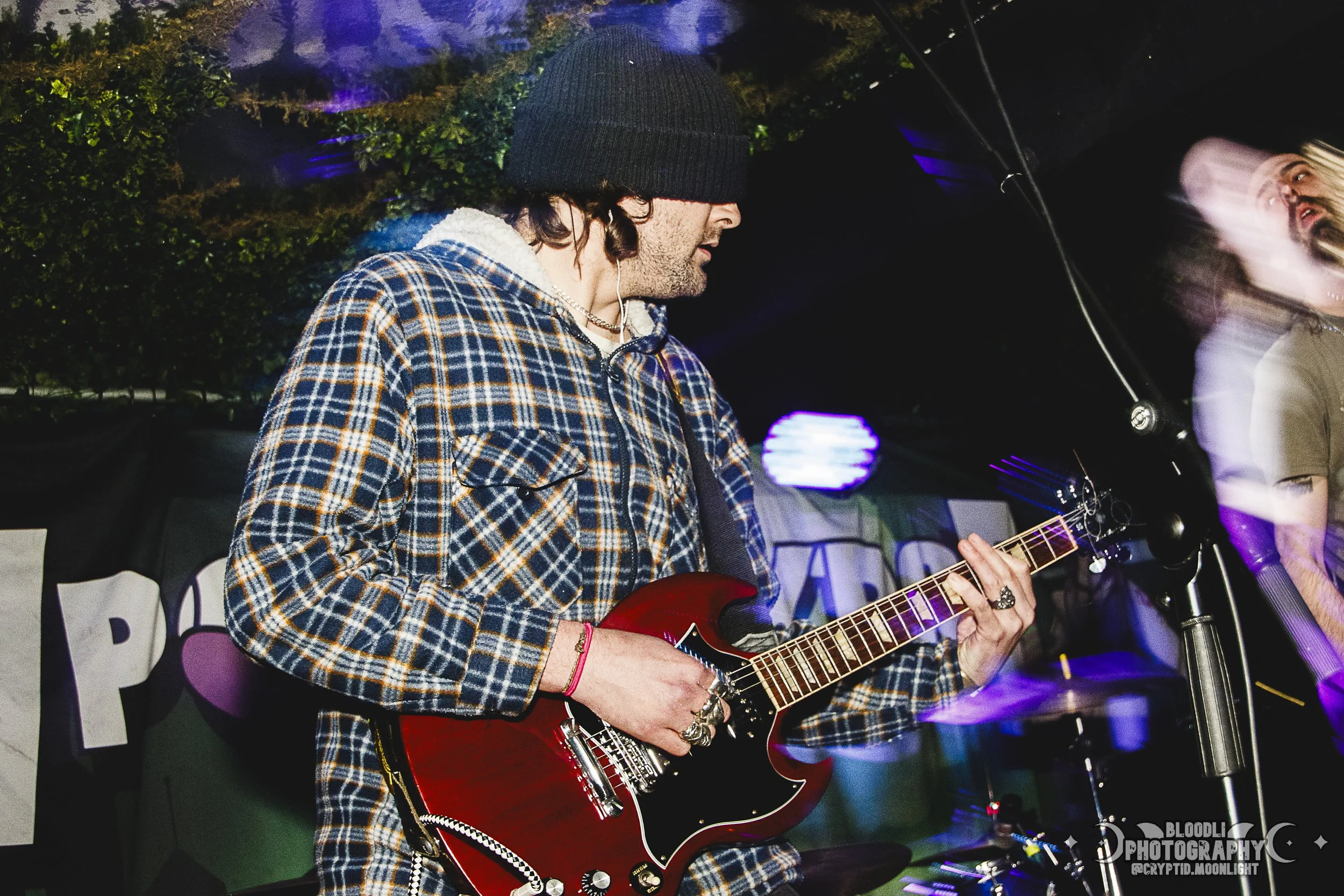 A person wearing a black beanie, plaid jacket, and rings plays a red electric guitar under stage lighting during a live music performance.