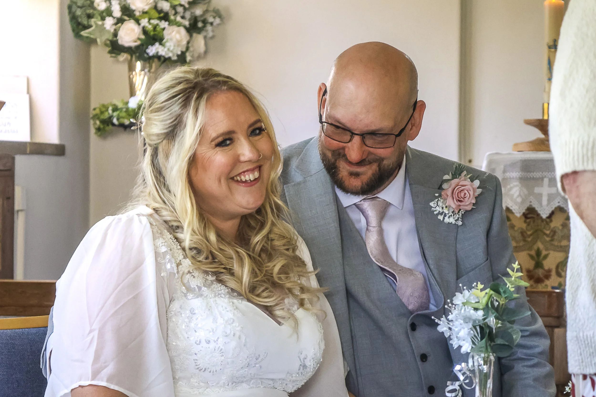 A smiling bride with long blonde hair and a groom with a beard and glasses all dressed in wedding attire inside a church, with a floral arrangement in front of them.