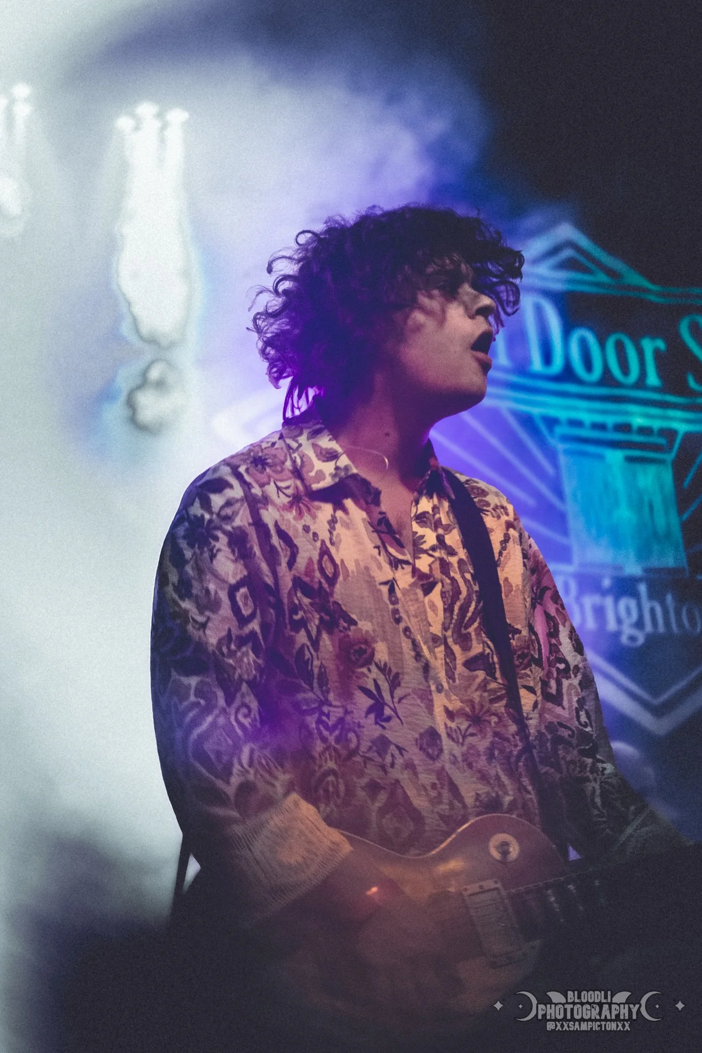 A person with curly hair playing an electric guitar at a concert, wearing a patterned shirt, with a neon sign in the background that reads 'Door S' and part of another word visible.